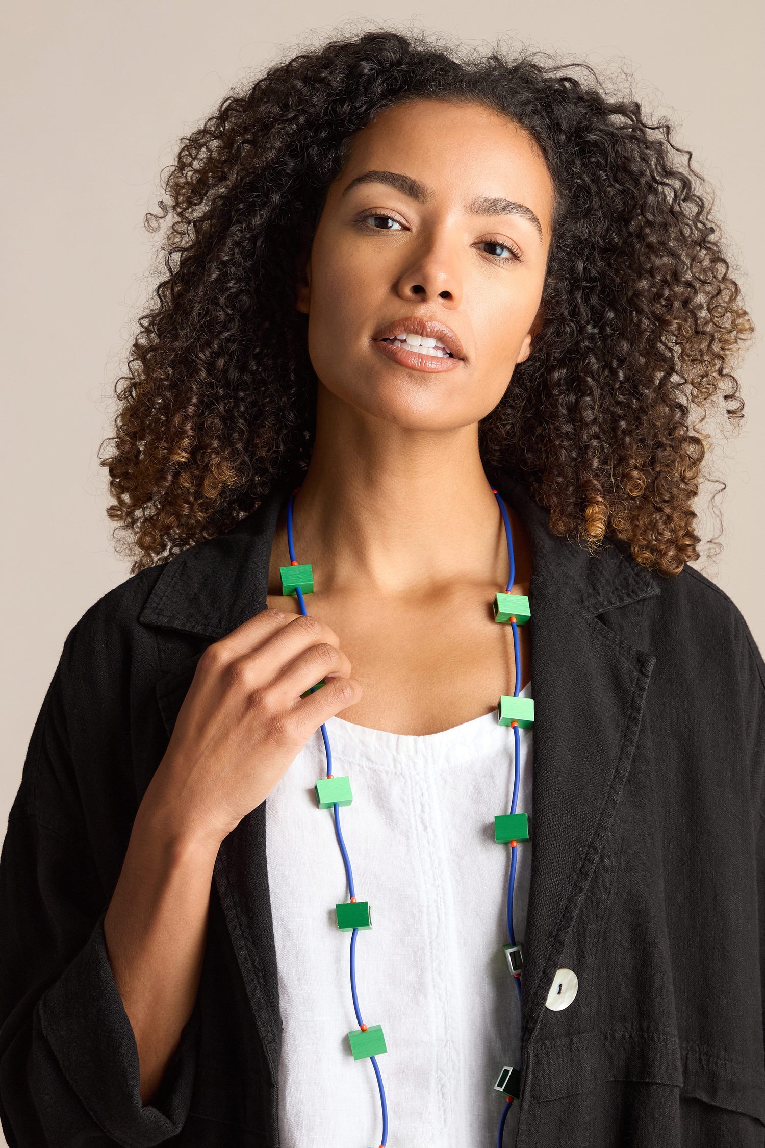 A woman with curly hair wearing a black jacket over a white top is adorned with a Long Aluminium Cube Necklace featuring green square-shaped beads by Chrisina Brampti, standing against a neutral background.