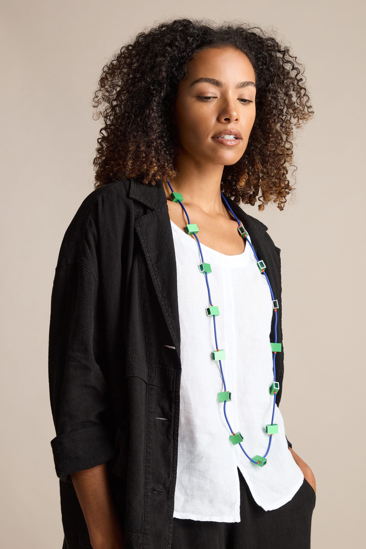 A person with curly hair, wearing a white top, black jacket, and a Long Aluminium Cube Necklace featuring blue and green accents, stands against a neutral background.