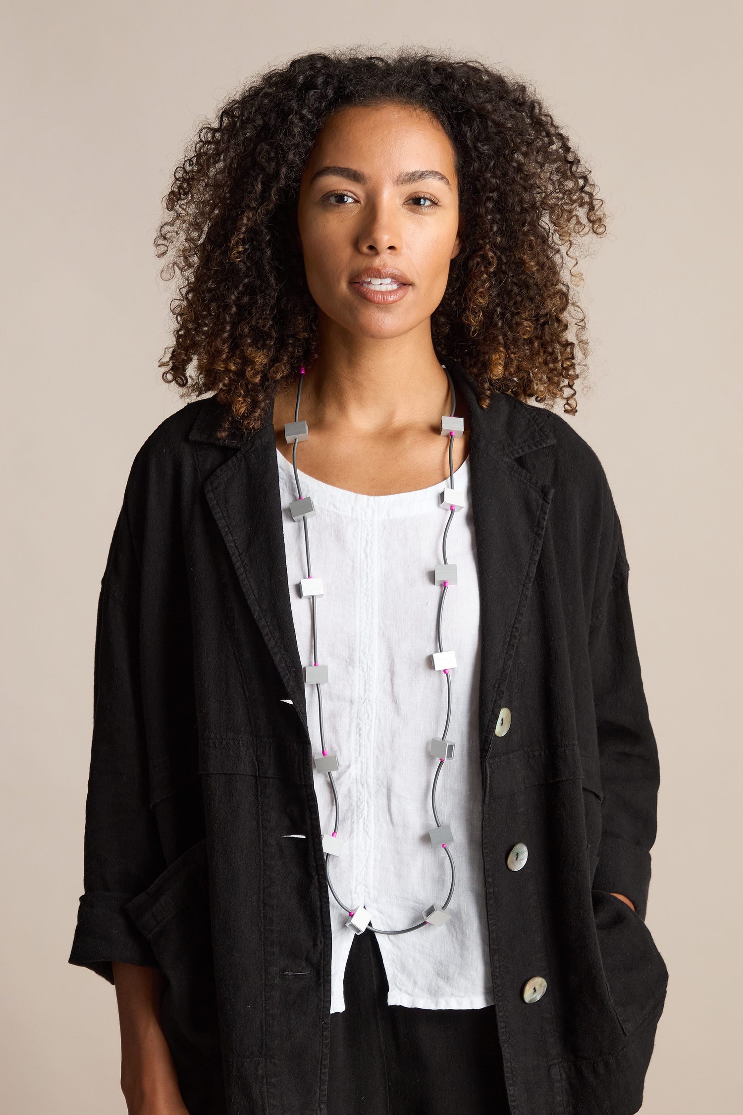 Person with curly hair wearing a black coat, white shirt, and a Long Aluminium Cube Necklace as a statement piece, standing against a neutral background.