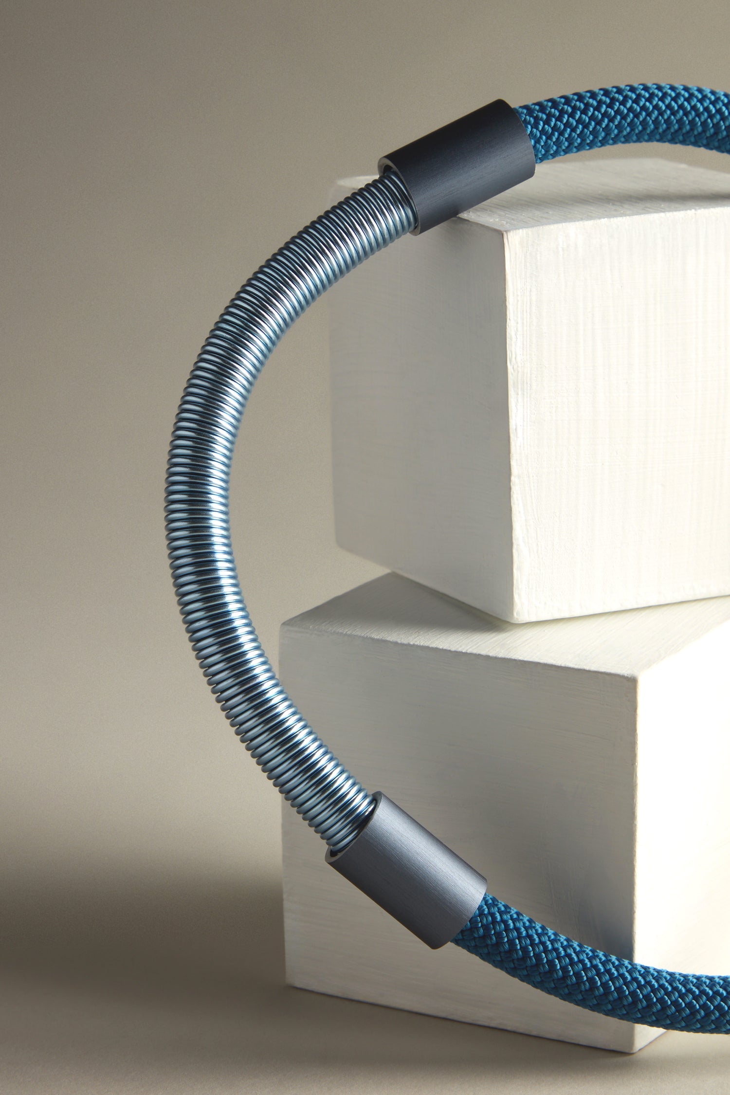 The Flow Cord Necklace showcases a flexible blue and silver cable with black connectors elegantly looped around off-white rectangular blocks, accented by durable anodized aluminum, and secured with a magnetic clasp.