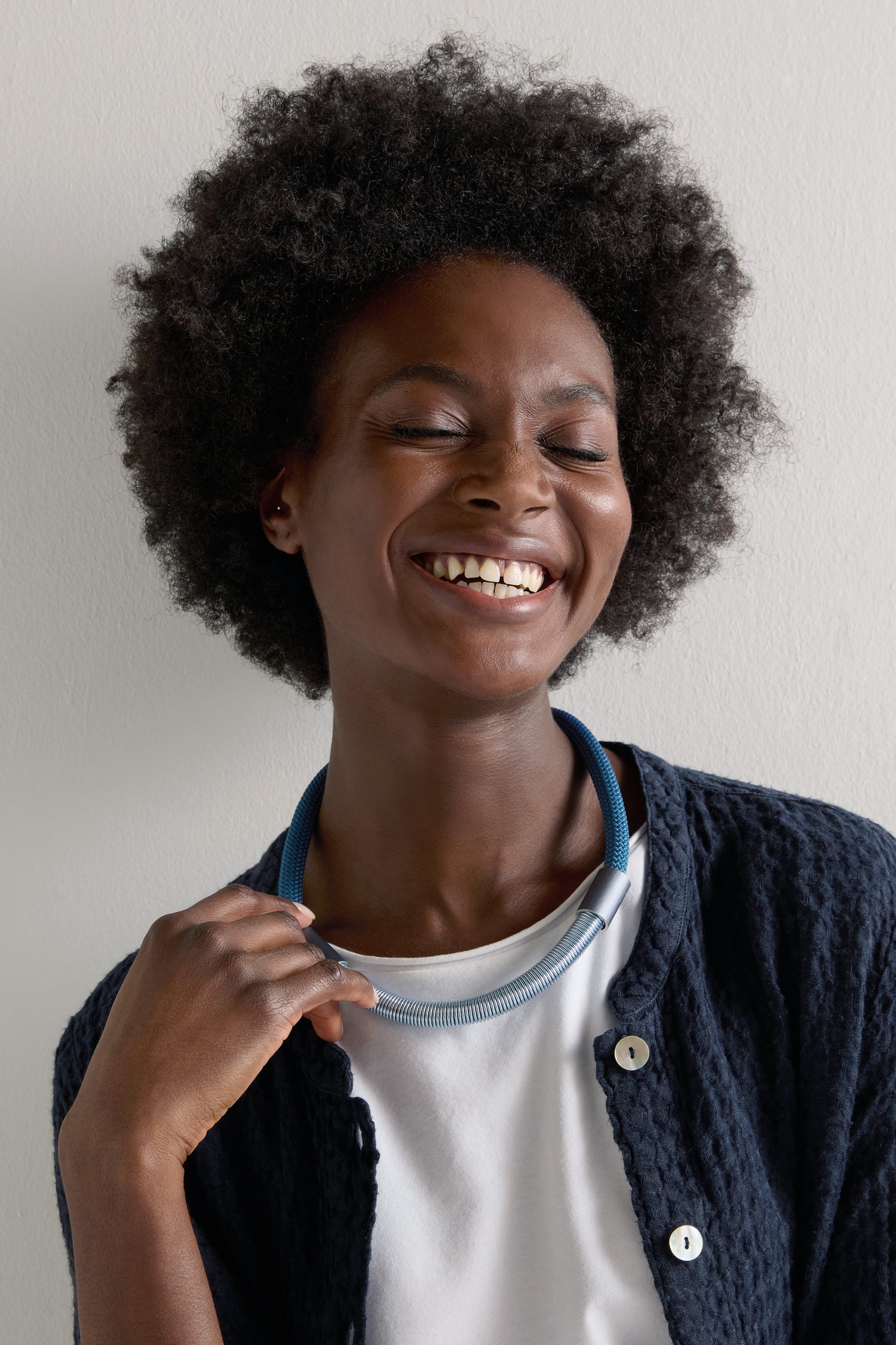 A curly-haired person in a navy cardigan and white shirt smiles with their eyes closed, wearing a bold Flow Cord Necklace crafted from anodized aluminum and fastened with a magnetic clasp.