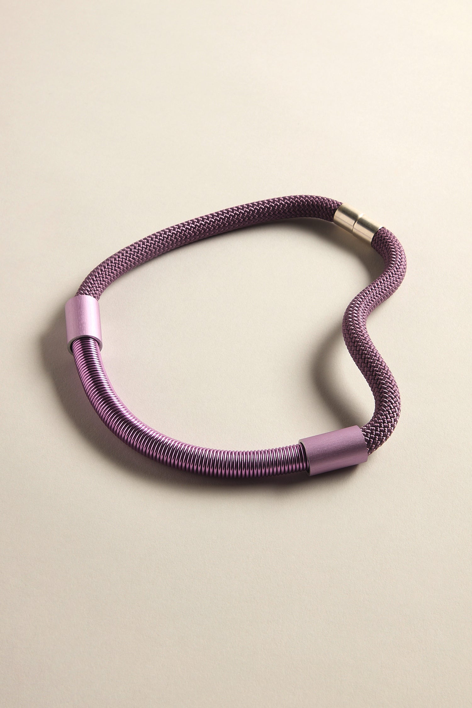 A purple Flow Cord Necklace with metallic connectors and a magnetic clasp is displayed against a light beige background.