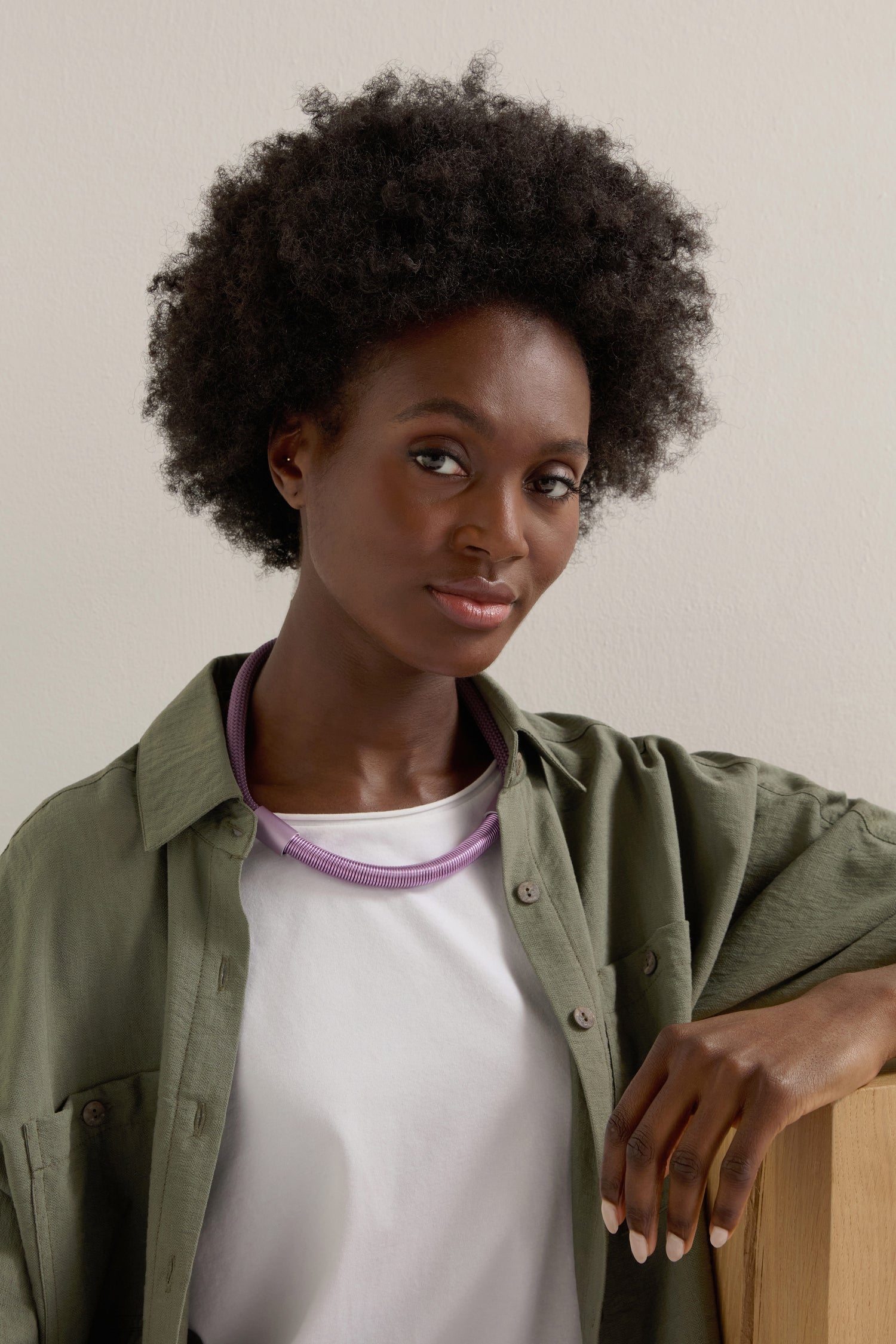 A woman with short curly hair in a green shirt over a white top displays her elegant Flow Cord Necklace. The necklace's purple anodized aluminum glistens as she leans against a wooden surface, highlighted by the off-white background, and it features a magnetic clasp.