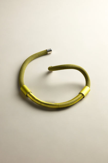 The Olive Flow Cord Necklace features a flexible metallic green design with anodised aluminium tubes, textured sections, and silver end caps, all highlighted by a magnetic clasp against a beige backdrop.