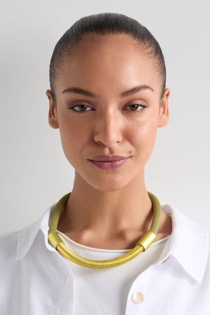 A person with a neat bun, wearing a white shirt and an Olive Flow Cord Necklace with an elegant gold anodised aluminum finish, smiles slightly against a plain background.