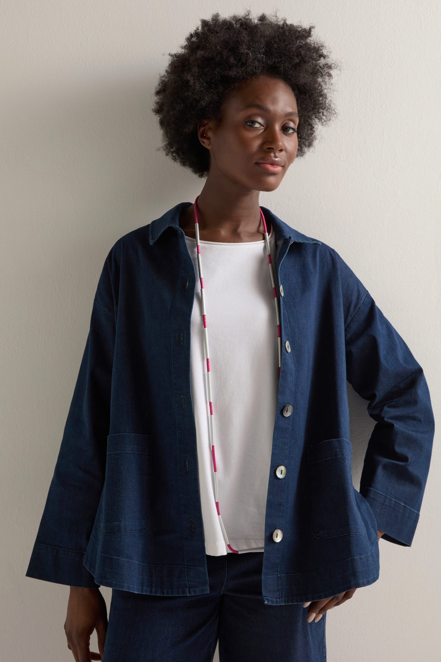 A person with curly hair wears a dark blue jacket over a white shirt, highlighted by the Long Cylinders Necklace in pink and white, showcasing bold simplicity against a plain background.