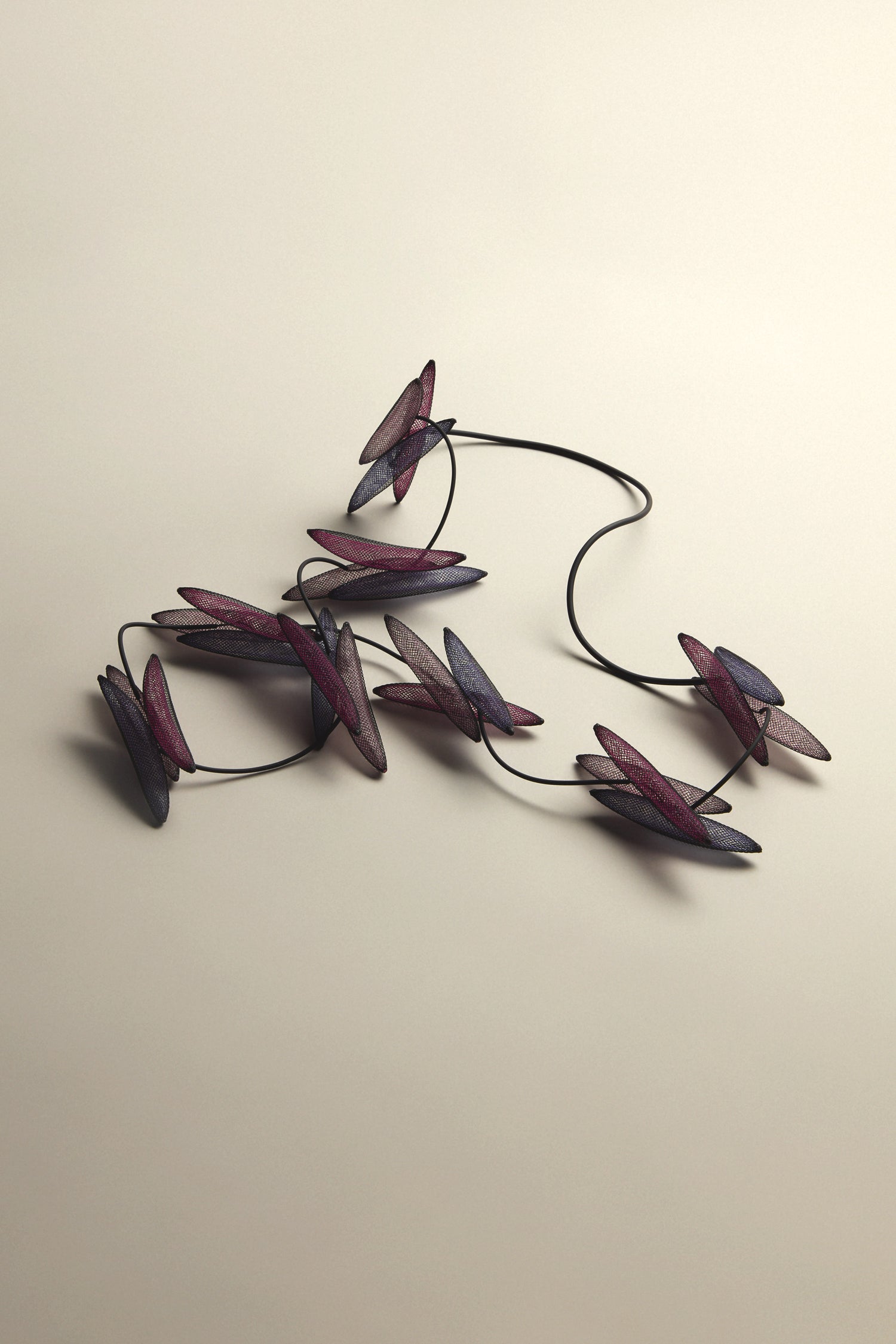 The Enchanted Necklace showcases red and purple leaf-shaped elements on a dark cord set against a light background, offering an ethereal, captivating statement piece with organic shapes.