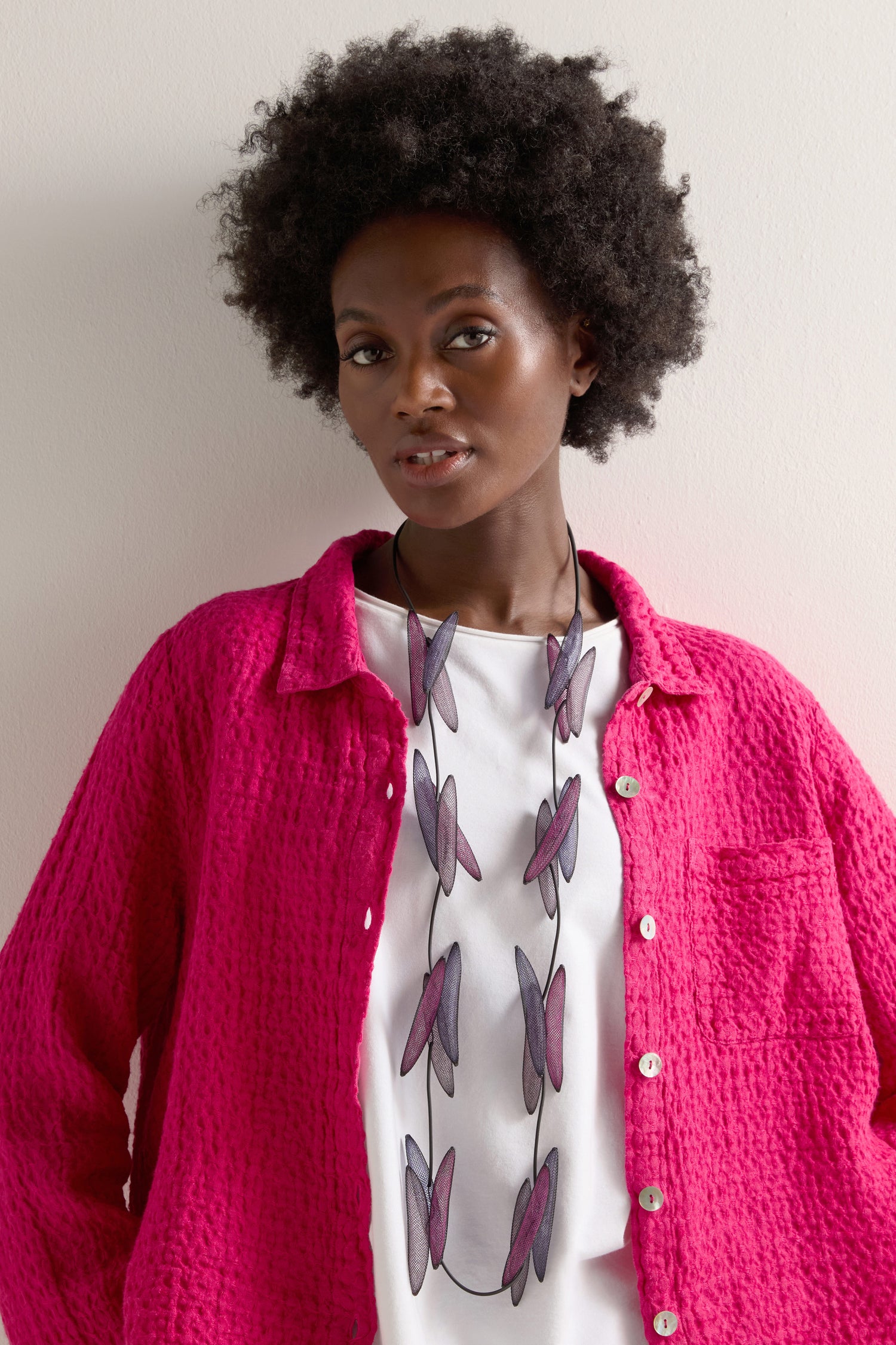 A woman with curly hair wears a pink textured jacket over a white shirt, beautifully accented by the Enchanted Necklace. This Christina Brampti piece features eye-catching purple and pink pendants, adding an elegant charm.