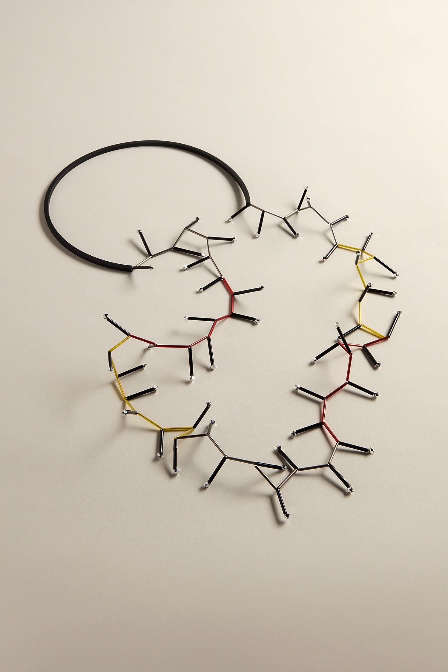 The Atomic Necklace is an avant-garde piece made from linked metal rods with red, yellow, and black details, creating an abstract shape on a light background—an unmistakable statement accessory.