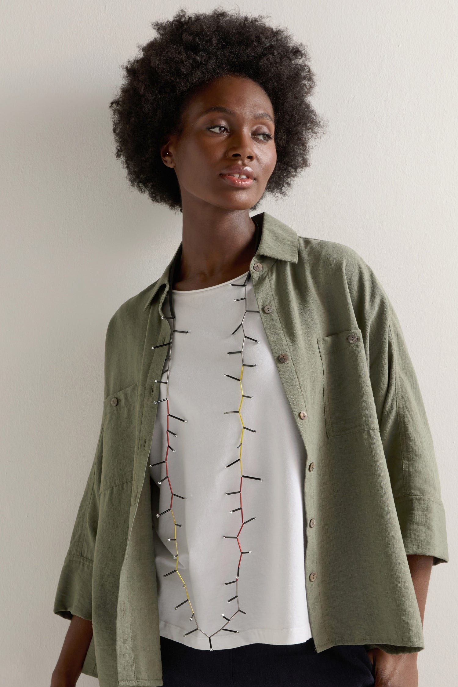 A person with curly hair wears a green button-up shirt over a white graphic T-shirt, complemented by the Atomic Necklace to make the look pop.