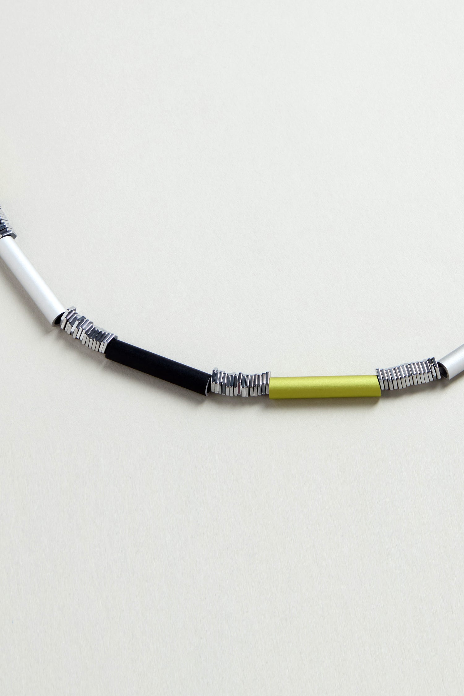 A close-up of the Atomic Silver Aluminium Hematite Necklace by Christina Brampti, showcasing alternating black, silver, white, and yellow cylindrical beads—an ideal choice for minimalist jewelry lovers.