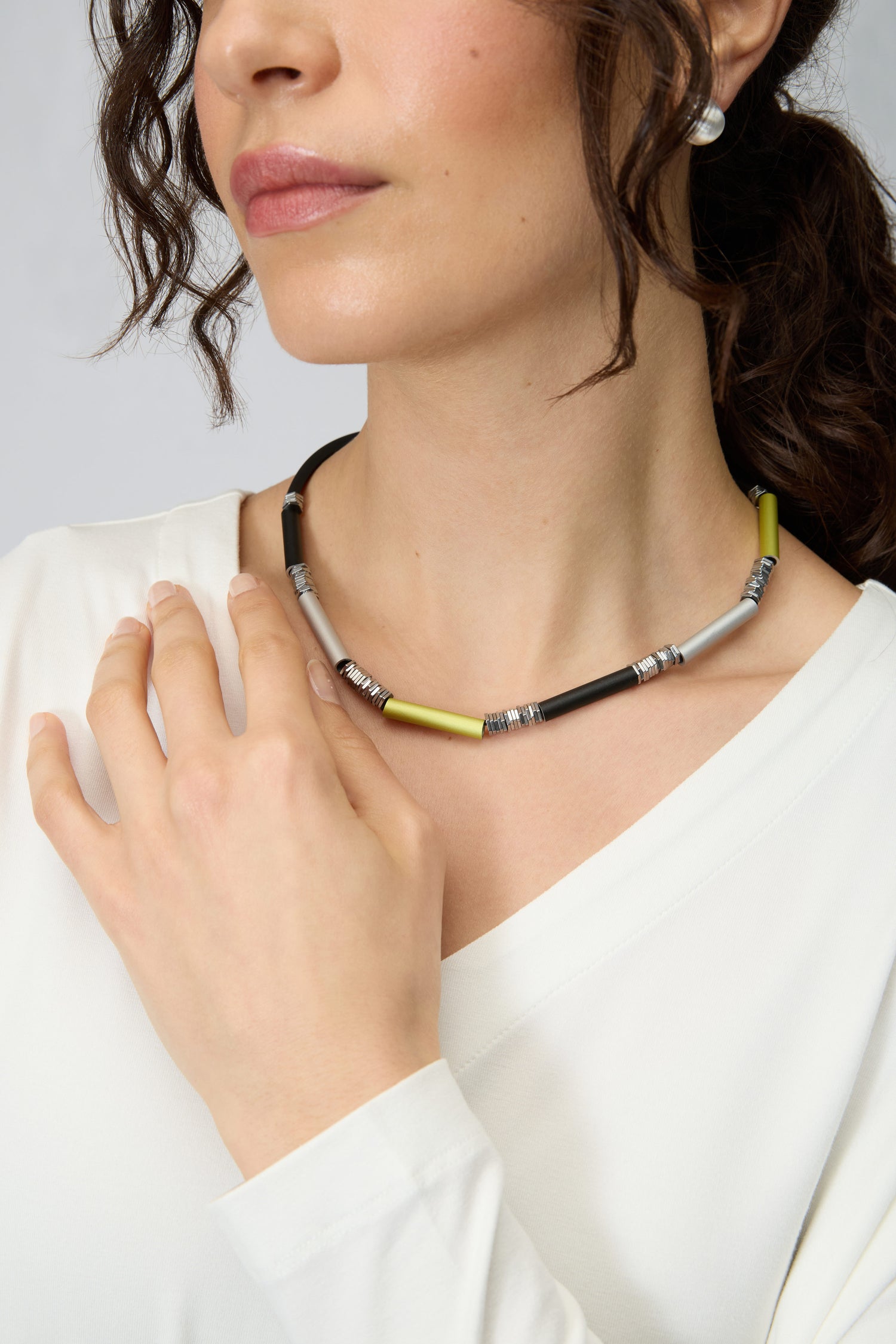 A woman wears a white top, silver stud earrings, and the Atomic Silver Aluminium Hematite Necklace by Christina Brampti, with one hand resting on her collarbone.
