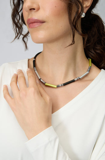A woman wears a white top, silver stud earrings, and the Atomic Silver Aluminium Hematite Necklace by Christina Brampti, with one hand resting on her collarbone.