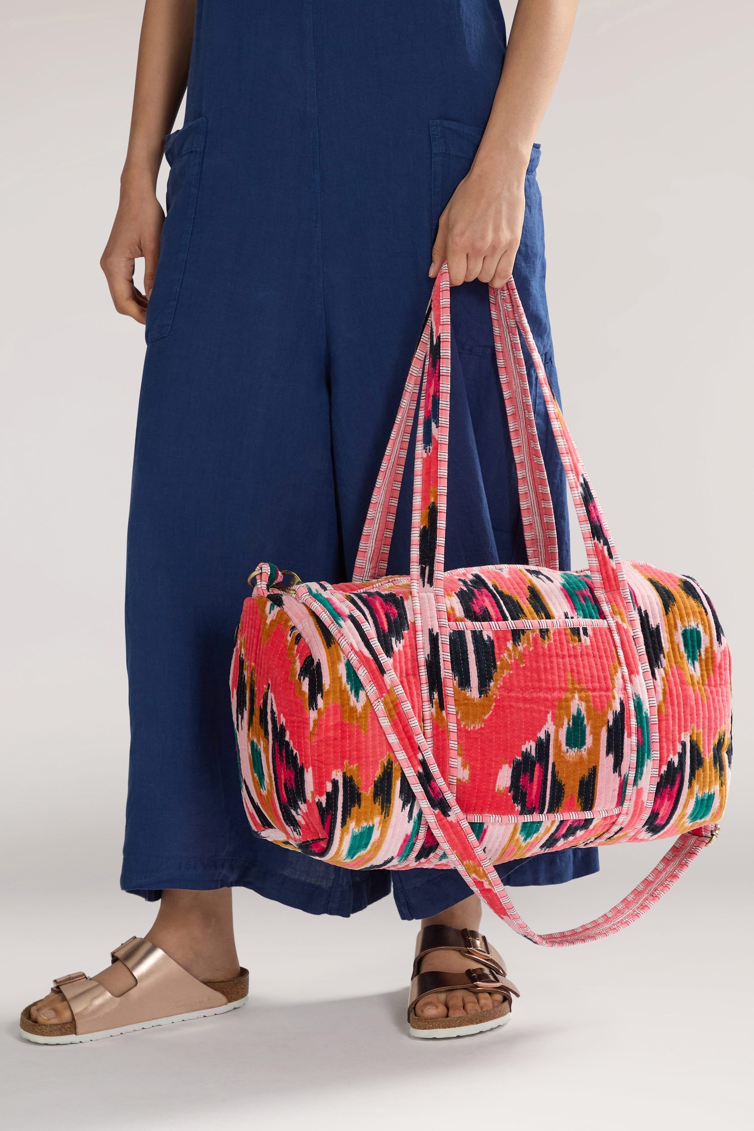 A person in a blue outfit holding an Ikat Velvet Weekend Bag, known for its resist-dyeing technique and colorful patterned design with pink handles. The person is wearing rose gold sandals.