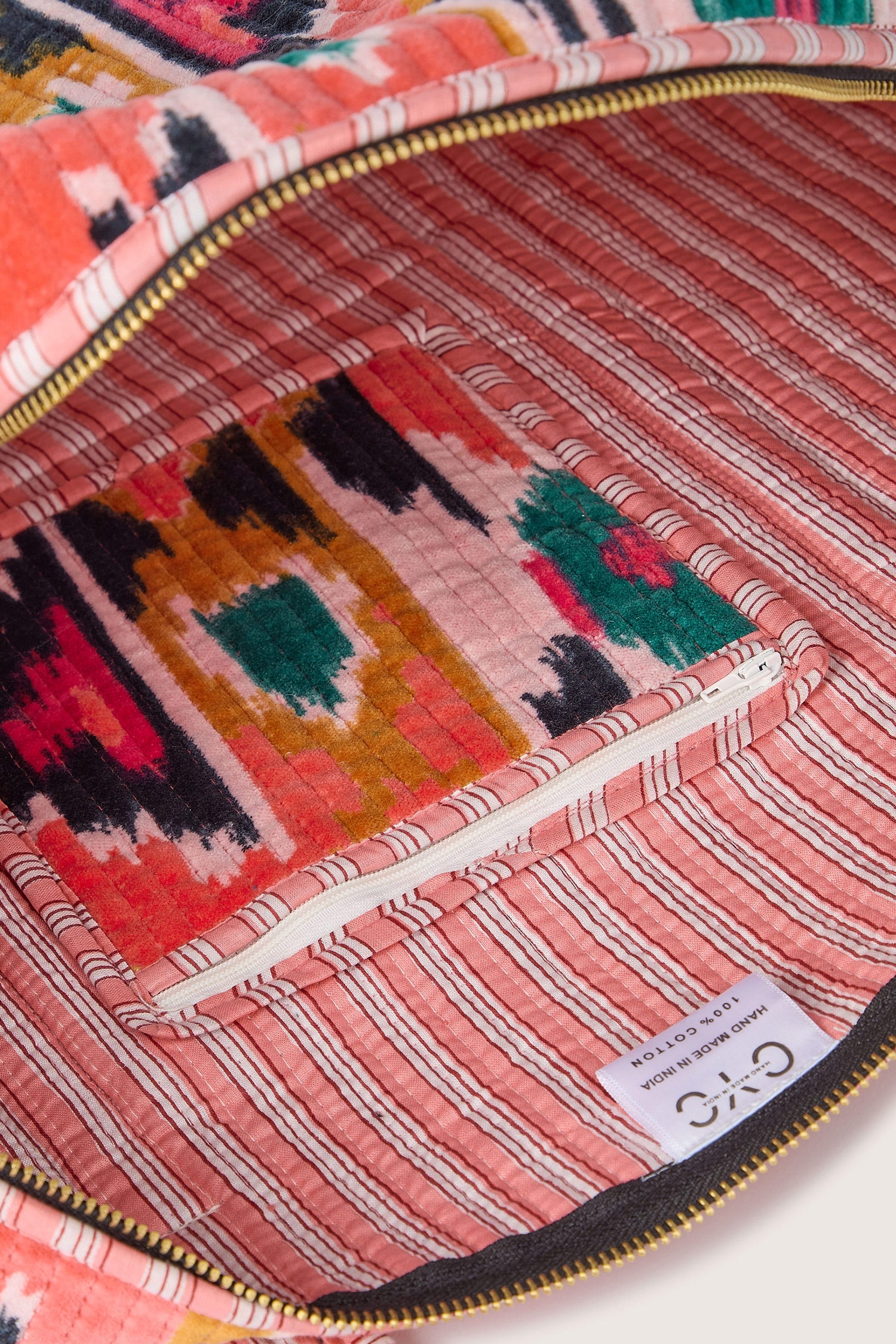 A brightly colored, patterned fabric-lined Ikat Velvet Weekend Bag is shown open, revealing its interior with a zippered pocket and a label that reads "Chico" and "Handcrafted." The versatile design makes it perfect for various occasions.