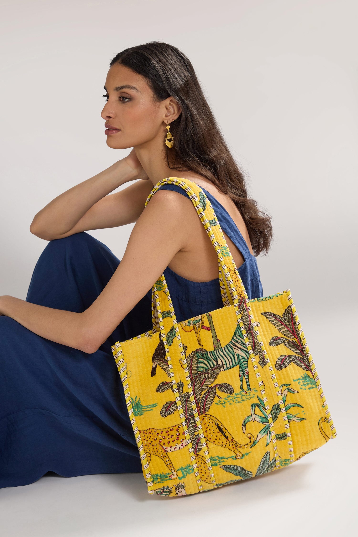 A woman in a blue dress sits with a Jungle Print Velvet Bag. She has long dark hair, gold earrings, and a focused expression. The background is plain and light-colored.