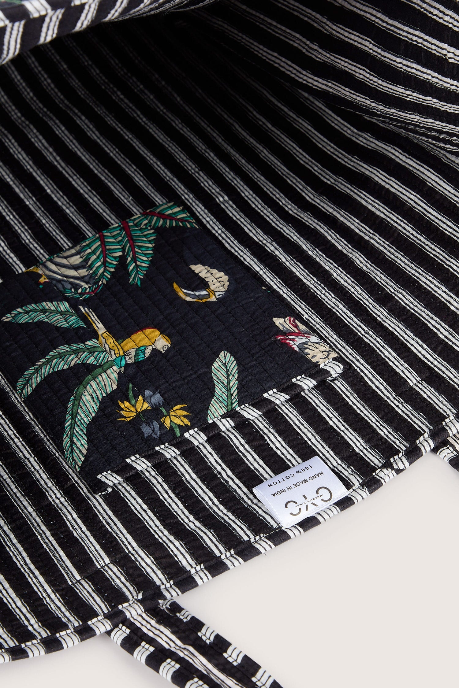 Close-up of a black and white striped quilted fabric featuring a small interior pocket with a colorful print of birds and foliage. A fabric label with the brand name is sewn inside, perfect for the Jungle Print Tote Bag.