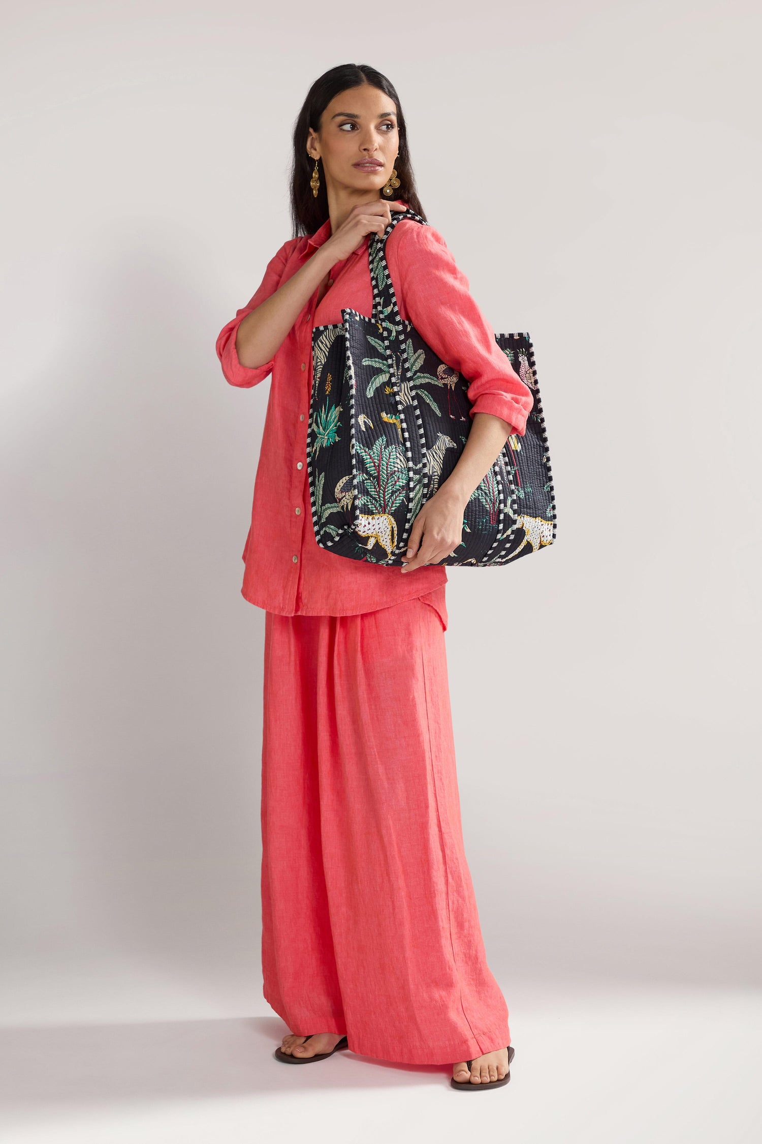 A woman stands against a neutral background, wearing a coral outfit and holding a large Jungle Print Tote Bag. She gazes off to the side and is wearing flat sandals.