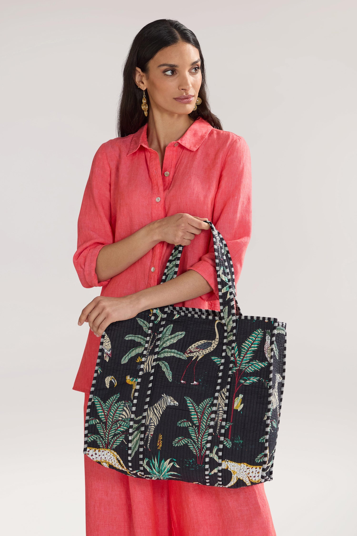 A woman in a coral-colored outfit holds a large black Jungle Print Tote Bag while looking to the side against a plain background.