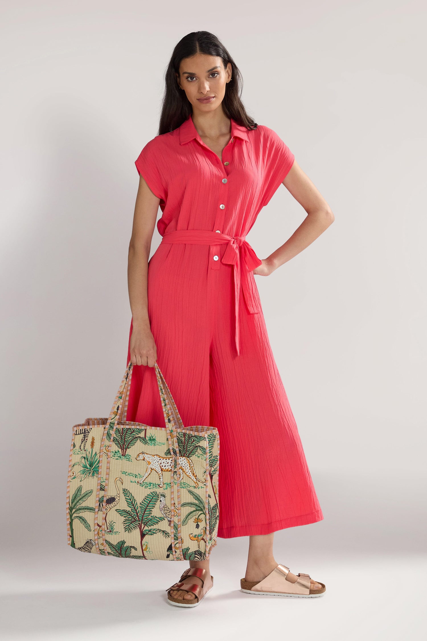 A woman in a bright coral jumpsuit with a tie waist stands confidently, holding a large Jungle Print Tote Bag and wearing tan sandals. She exudes effortless style, even carrying her essentials in quilted fabric for added elegance.