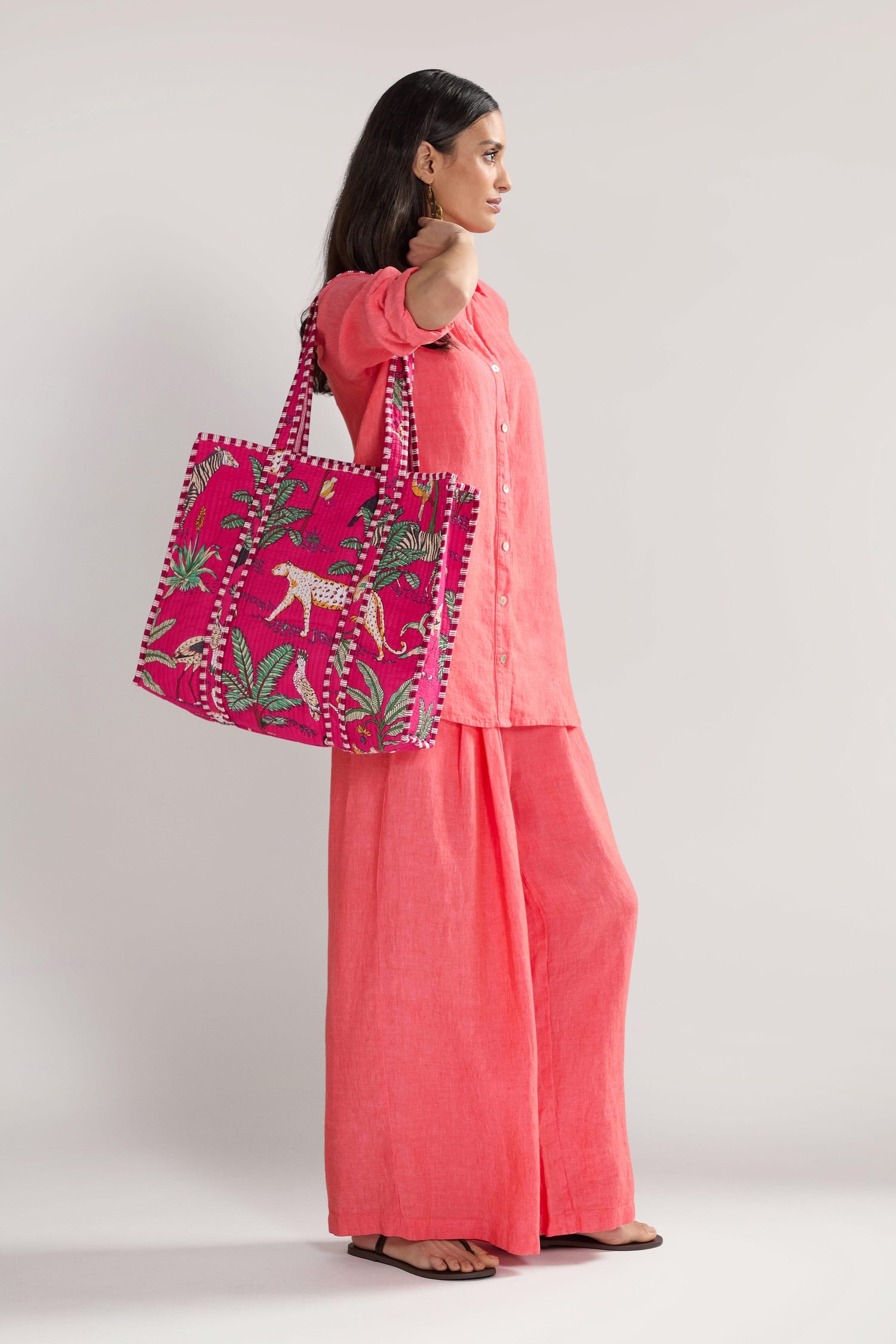 A woman stands sideways wearing a coral-colored outfit and holding a large pink Jungle Print Tote Bag from the Conscious Yoga Collective, adorned with a colorful tropical print featuring a white tiger and green leaves.