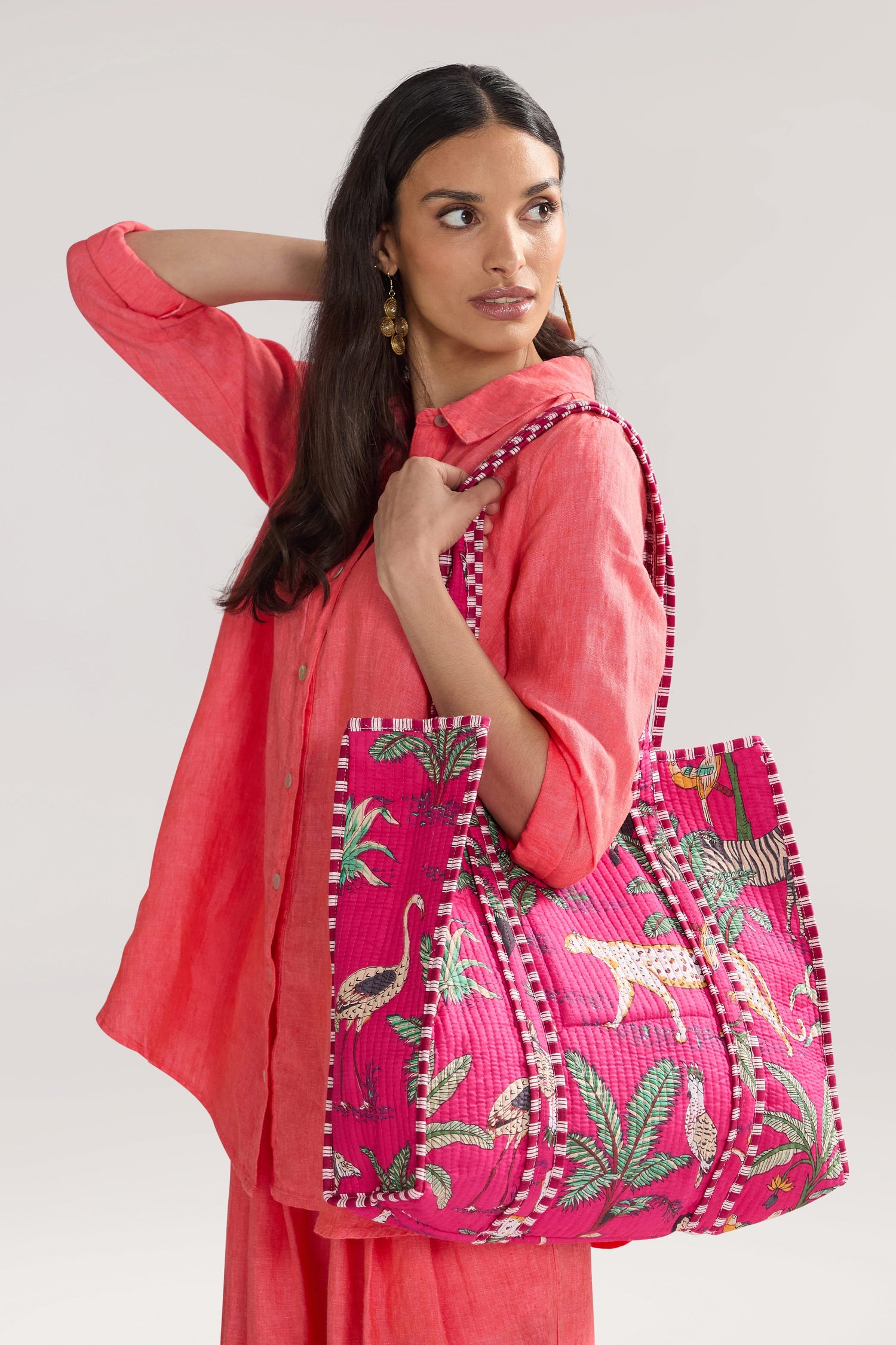 A woman wearing a pink outfit holds a large, pink-patterned Jungle Print Tote Bag with tropical animal designs made from quilted fabric. She looks to the side with her other hand raised to her head, exuding the serene confidence of someone from the Conscious Yoga Collective.