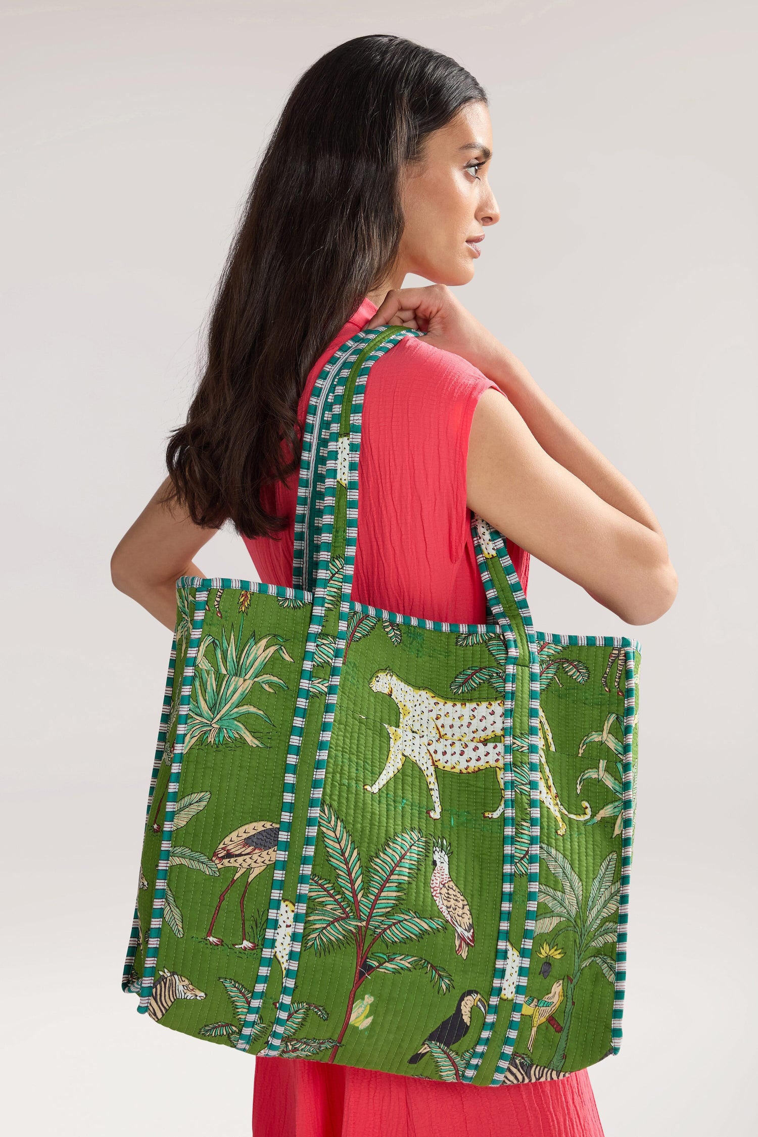 Person with long dark hair wearing a bright pink outfit and carrying a large green Jungle Print Tote Bag made of quilted fabric with blue-striped handles.