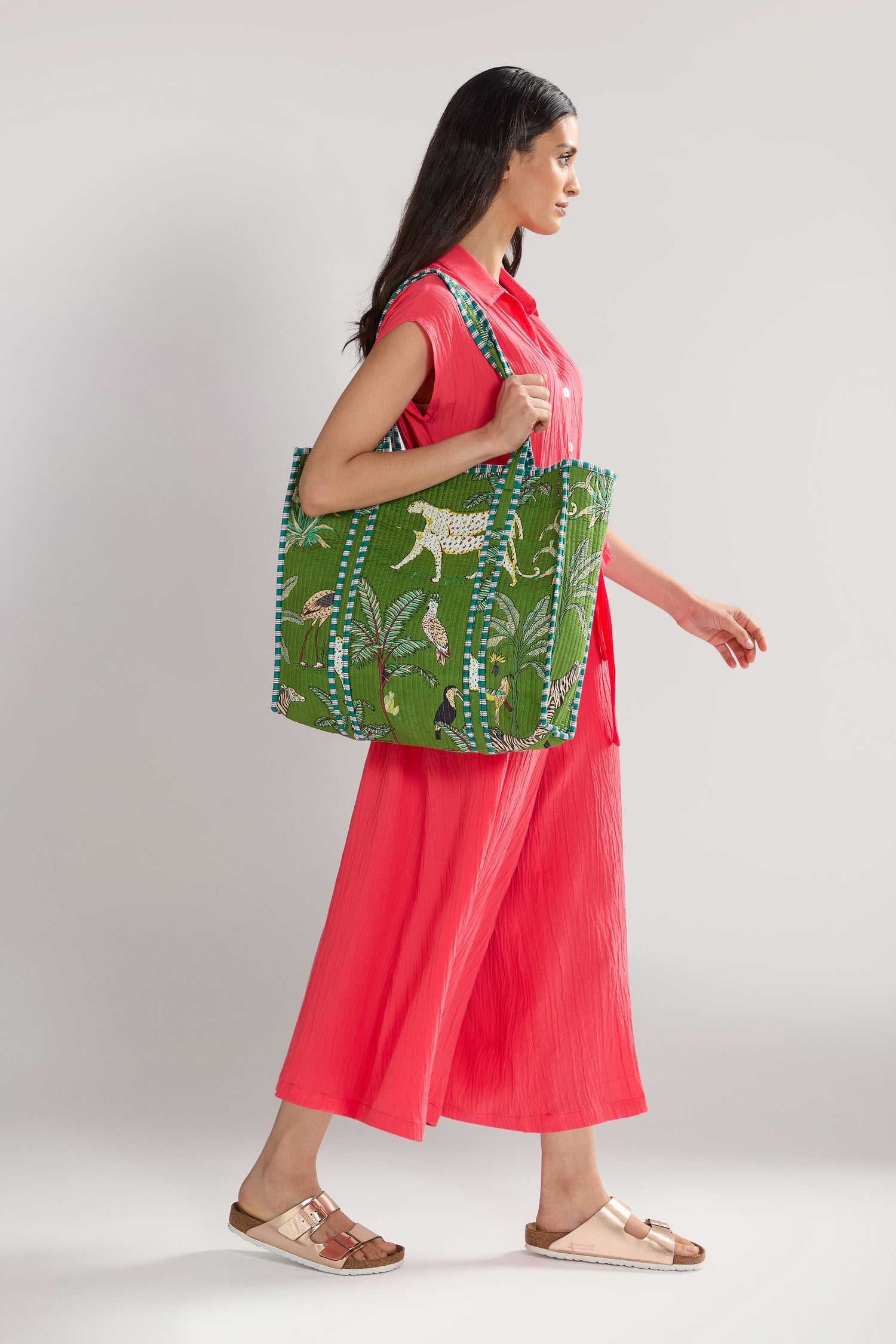 A woman in a pink outfit walks while holding a large green Jungle Print Tote Bag with an animal and plant design. She is wearing beige sandals.