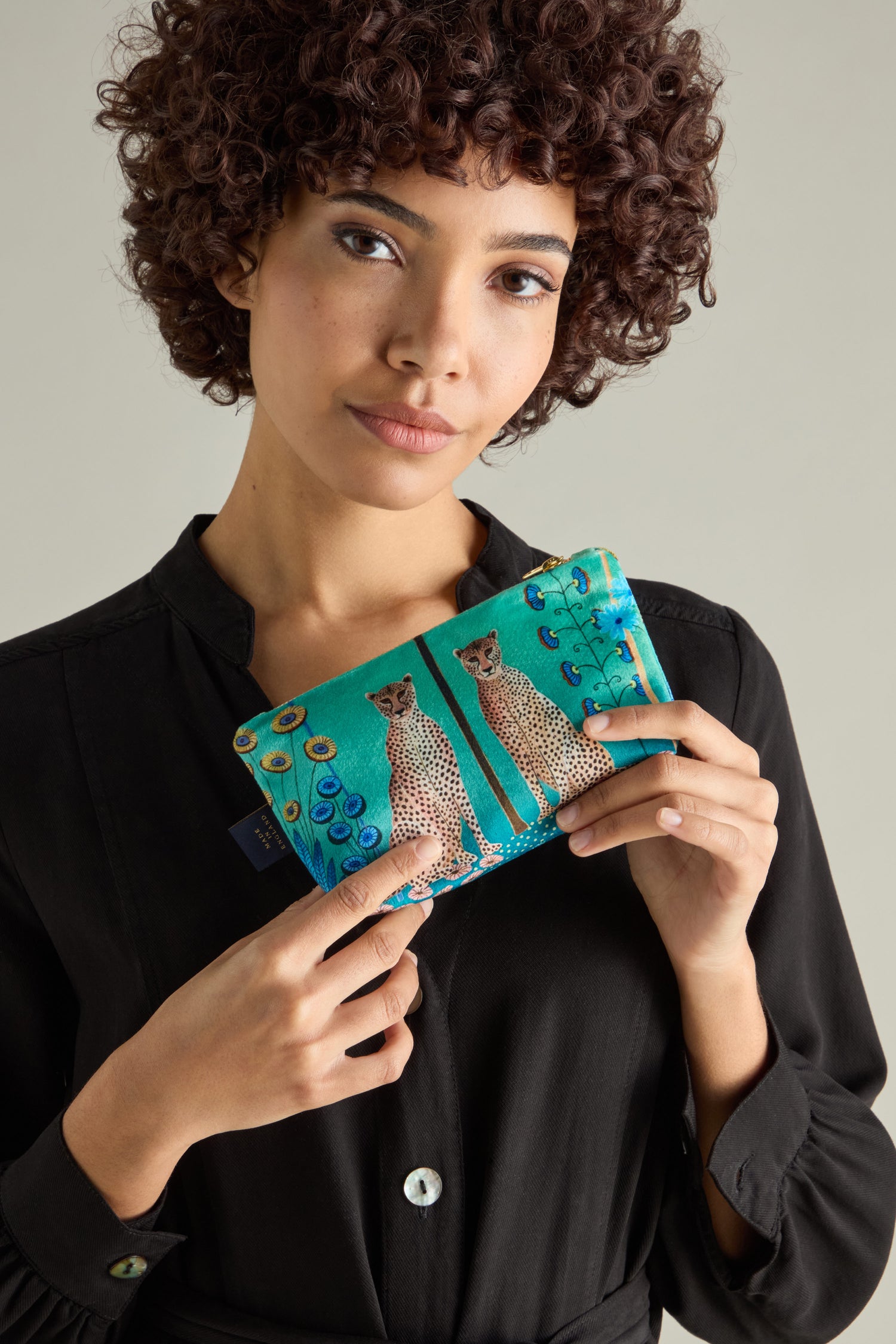 With curly hair, a person holds the Small Sunshine Through My Window Velvet Bag in turquoise, featuring cheetah designs and peacock motifs, while wearing a black button-up shirt. The scene captures the essence of wild elegance.