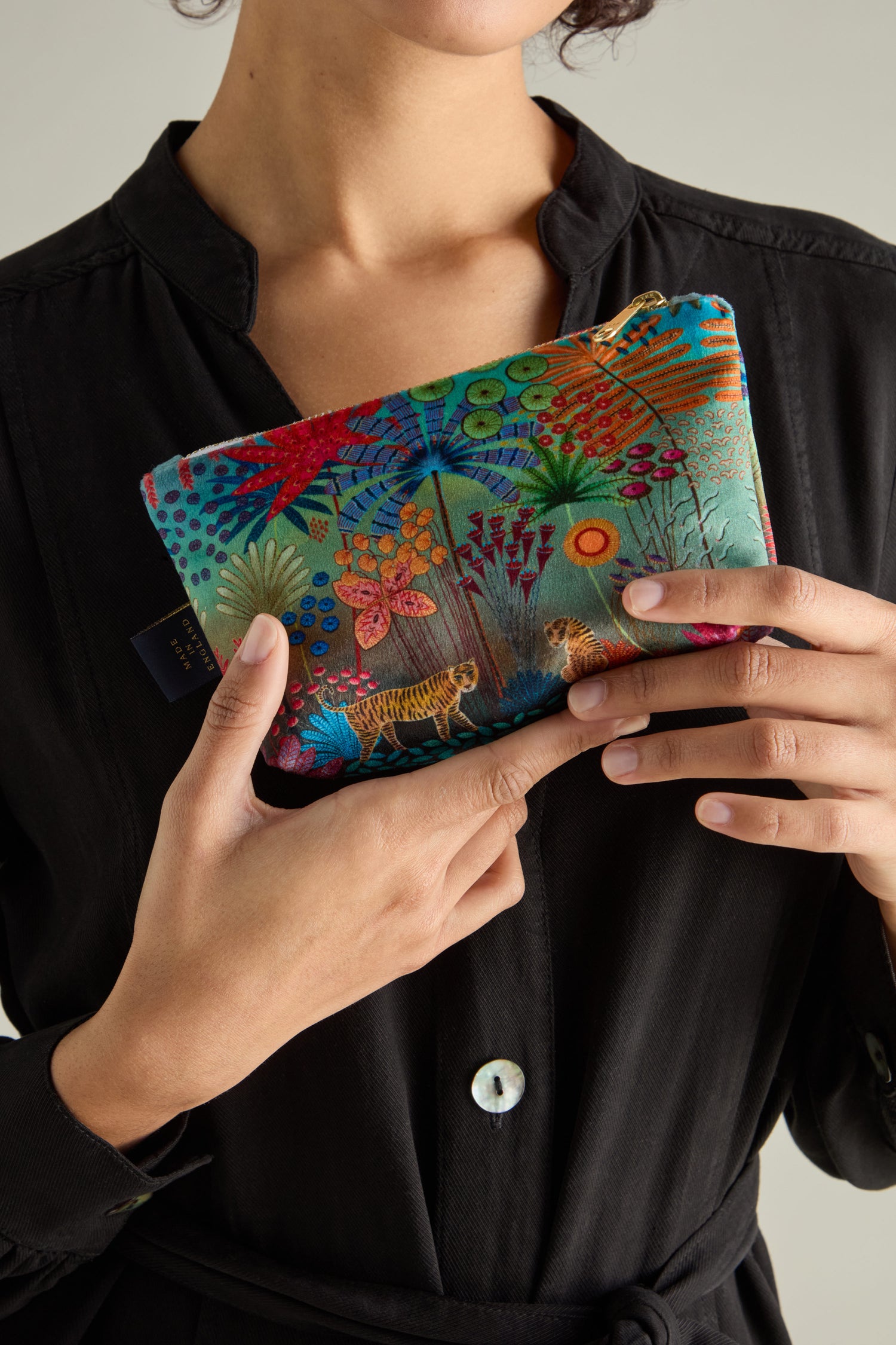 A person holds a vibrant Small Tale of Two Tigers Velvet Bag with a jungle design, set against a black button-up shirt.