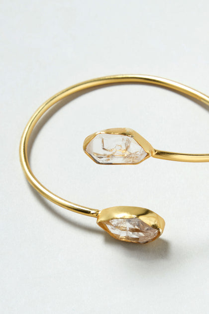 The Gold Plated Quartz Herkimer Diamond Bracelet is an 18k gold plated open cuff bangle with two irregular clear stones at each end, shown on a light background.