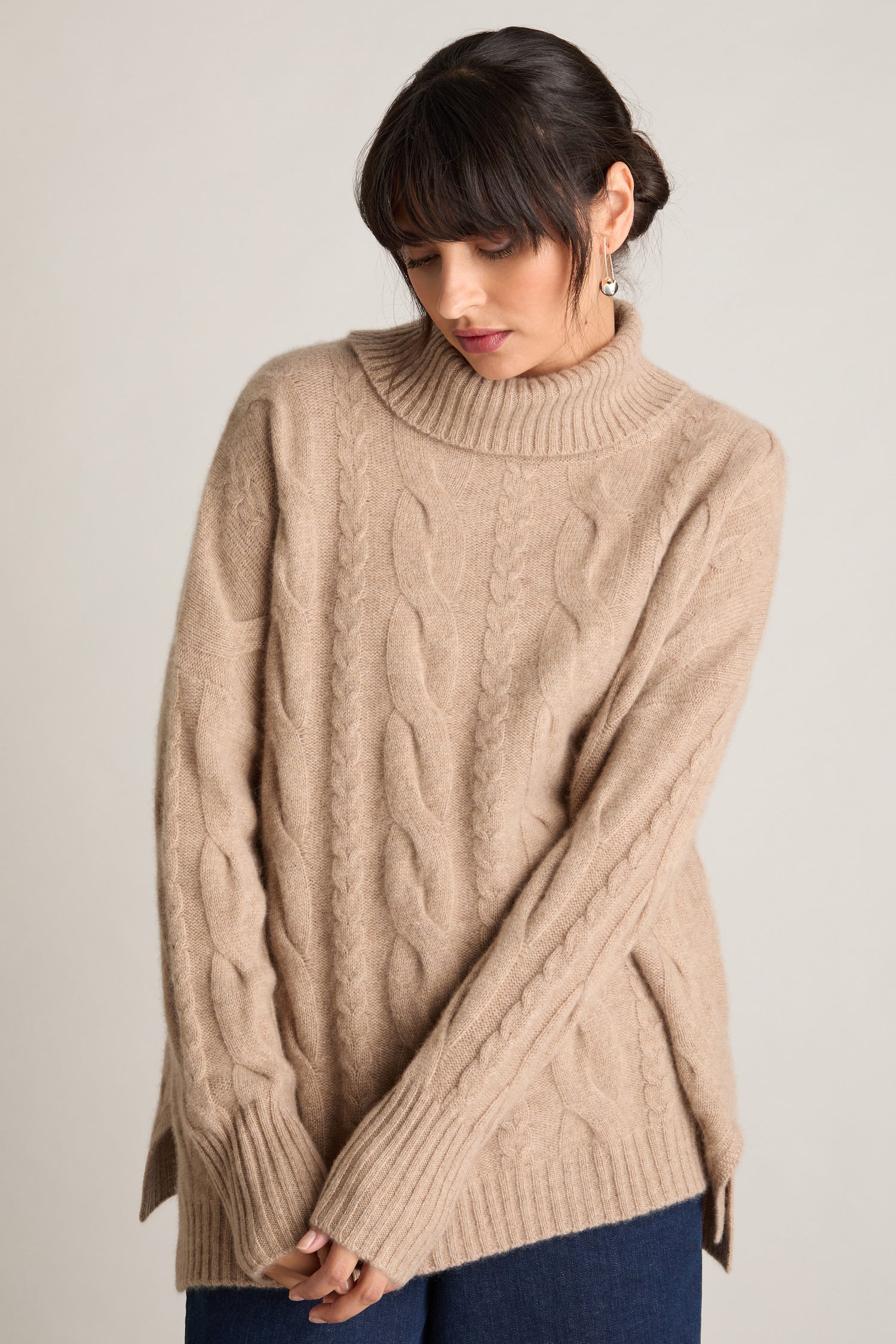 A woman wearing a Loose Cable Cashmere Knit sweater in beige, featuring long sleeves and a turtleneck design, stands against a plain background. The relaxed fit of her sweater perfectly complements her dark pants for a casual yet stylish look.