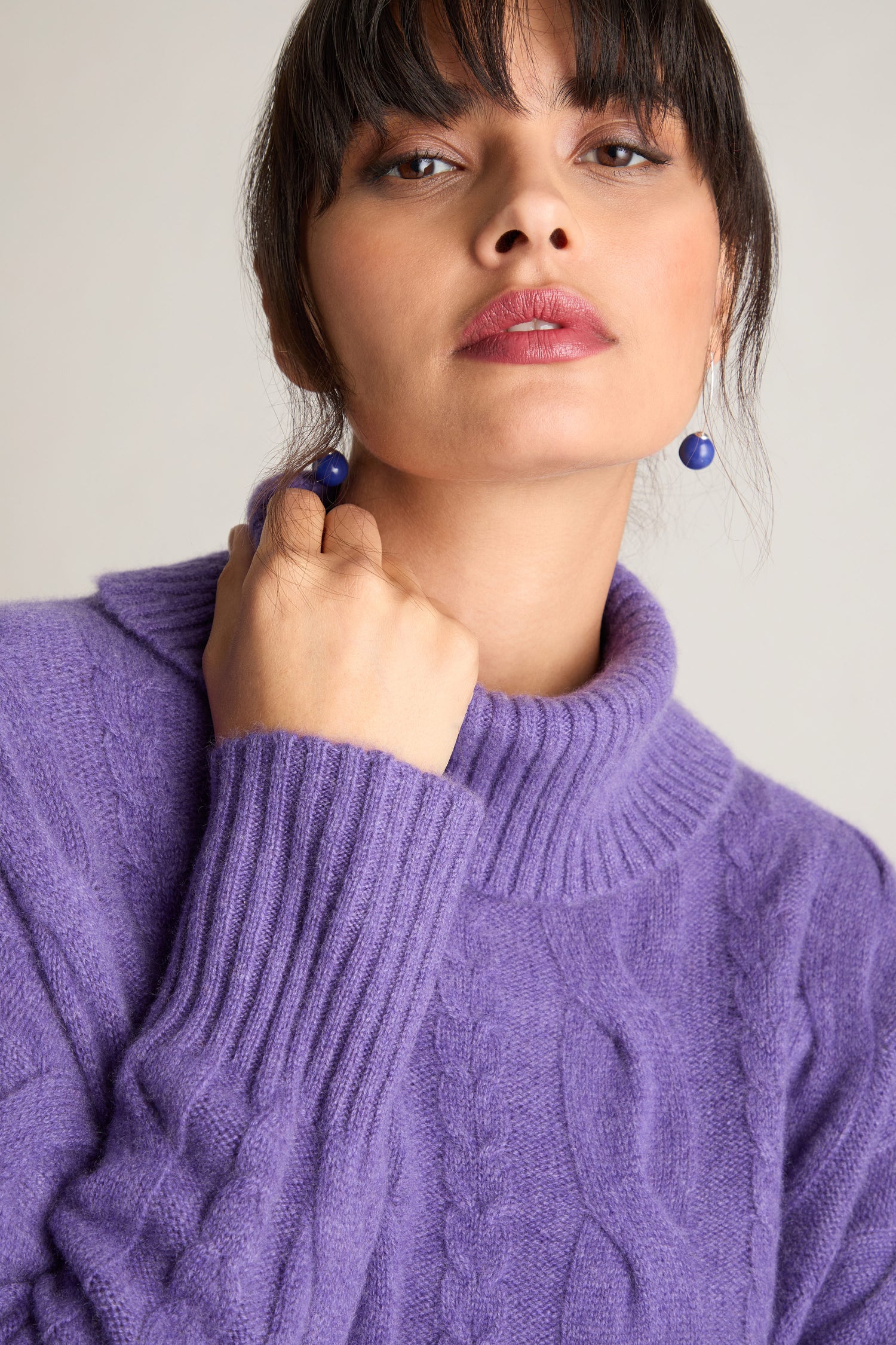 A woman wearing a Loose Cable Cashmere Knit in purple and blue earrings looks directly at the camera with a neutral expression.