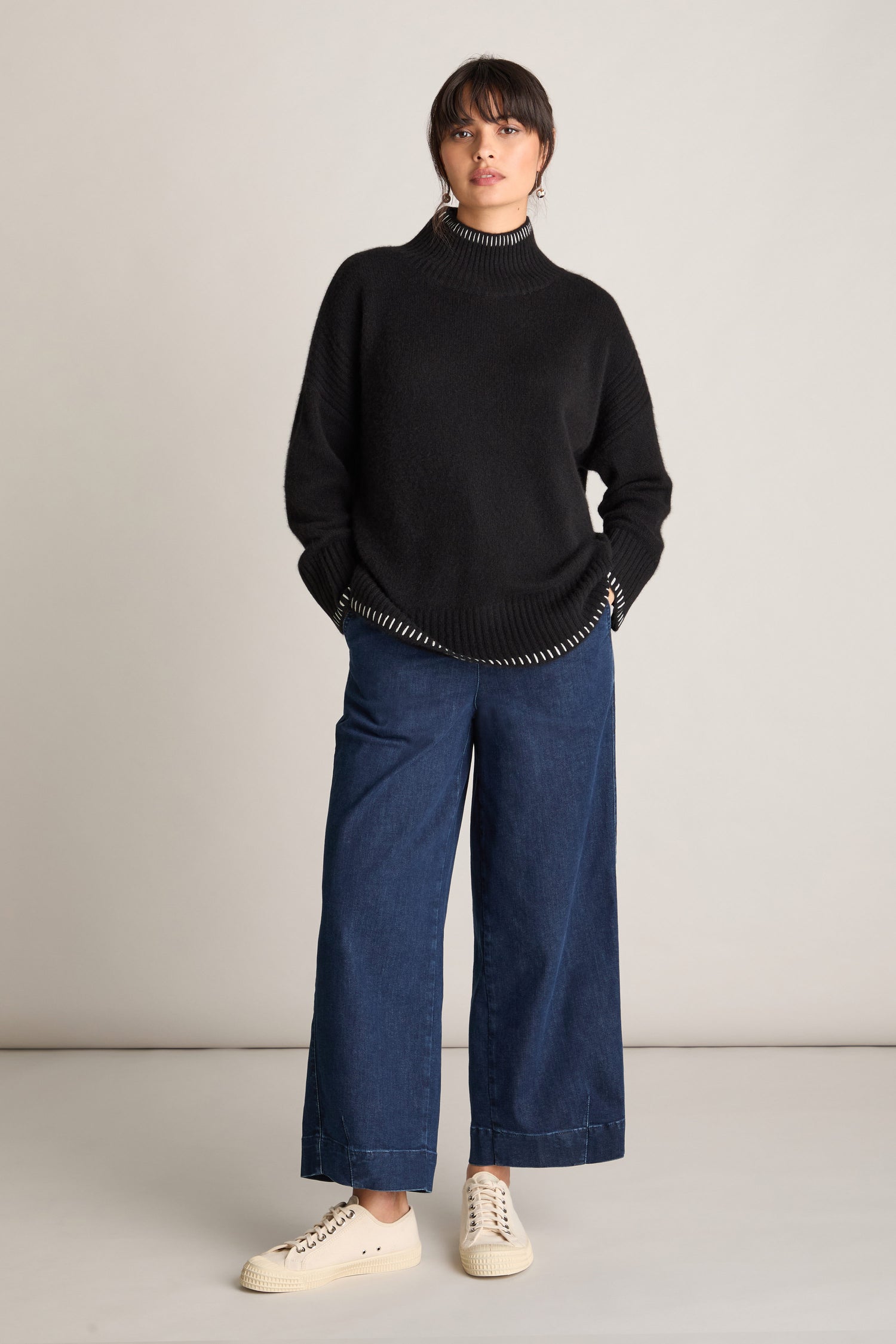 An individual stands against a plain background, wearing an Estheme Blanket Stitch Funnel Neck Cashmere Knit sweater, blue wide-leg jeans, and white sneakers with their hands in their pockets.