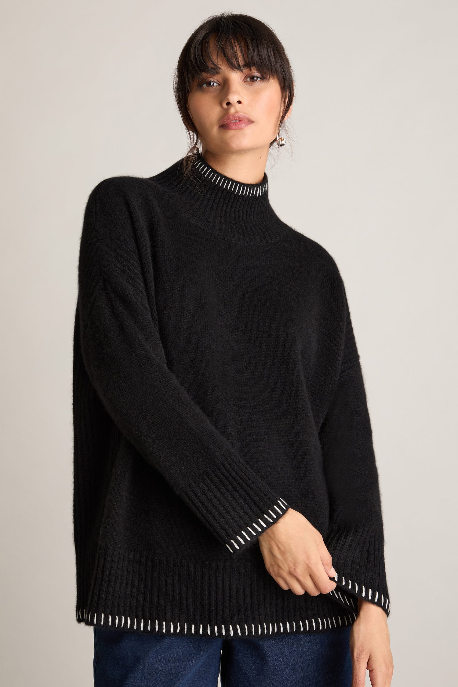 A woman with dark hair and bangs is wearing the Blanket Stitch Funnel Neck Cashmere Knit in black, featuring a white trim, as she stands against a plain background.