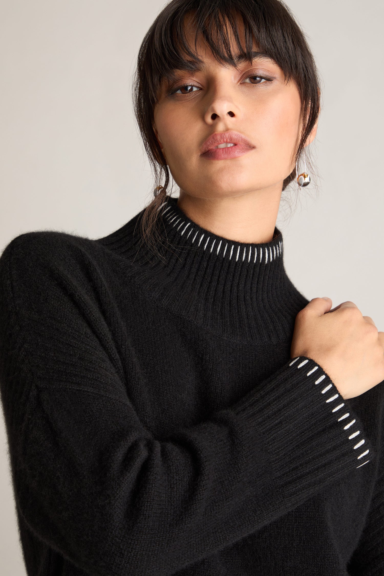 A person with dark hair and bangs is wearing a black Blanket Stitch Funnel Neck Cashmere Knit, posing with one hand touching the opposite shoulder.