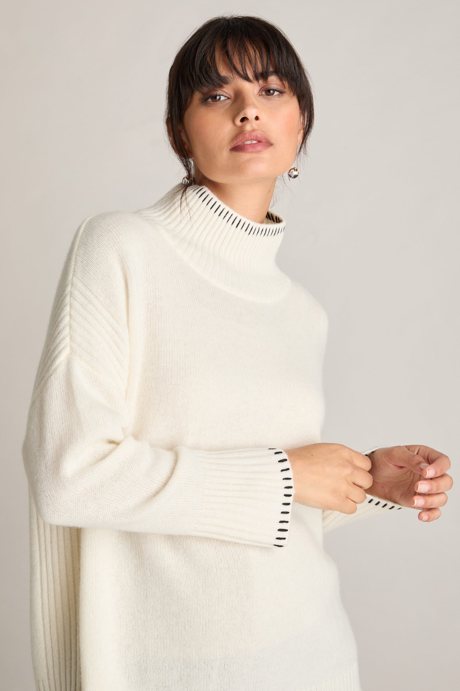 A person wearing a Blanket Stitch Funnel Neck Cashmere Knit in white with black striped edges stands against a plain background, showcasing the elegance of cashmere knit.