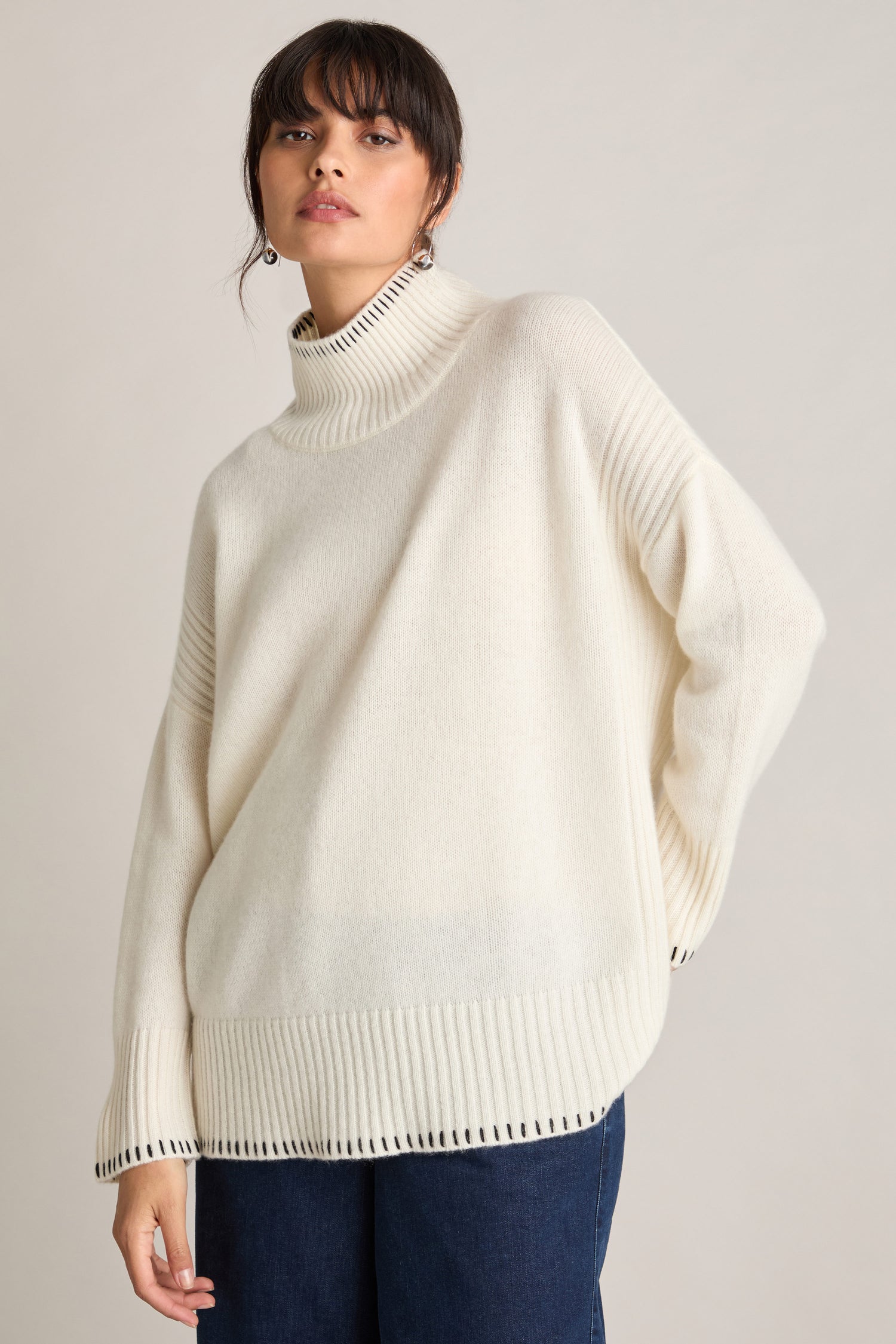 A person with dark hair stands wearing a loose, off-white Blanket Stitch Funnel Neck Cashmere Knit sweater and blue jeans. The background is plain and light-colored.