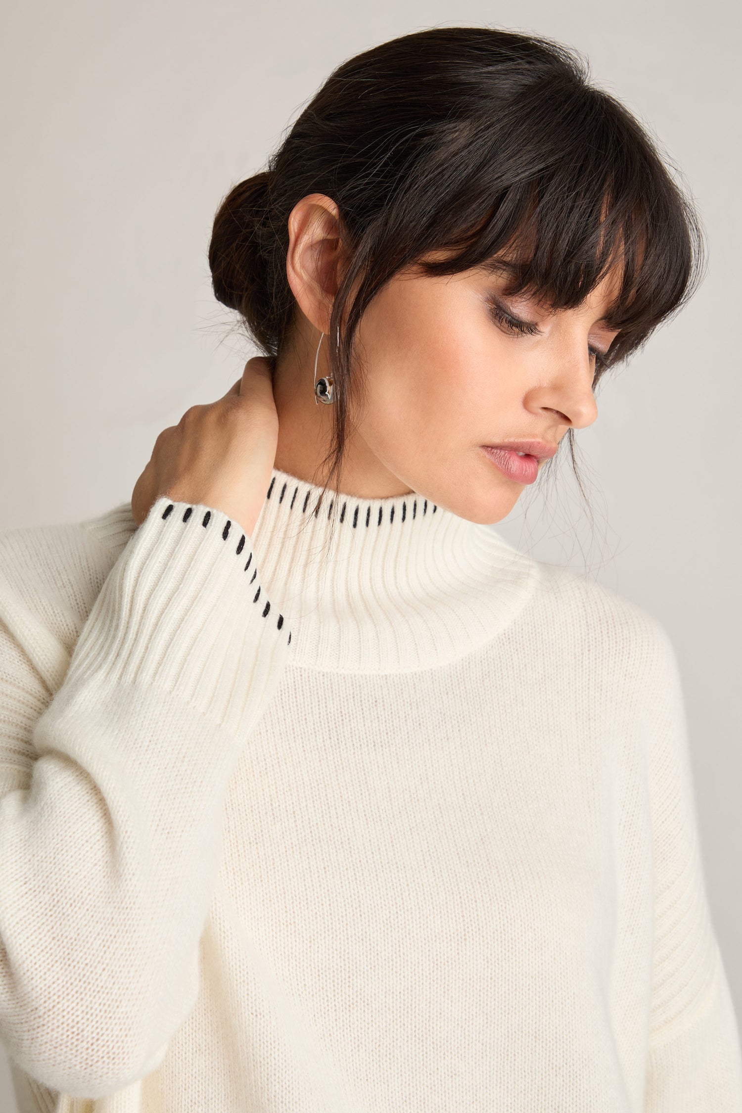 A person with dark hair styled in a low bun, wearing a Blanket Stitch Funnel Neck Cashmere Knit in white with black detailing, looks down thoughtfully with one hand touching their neck.