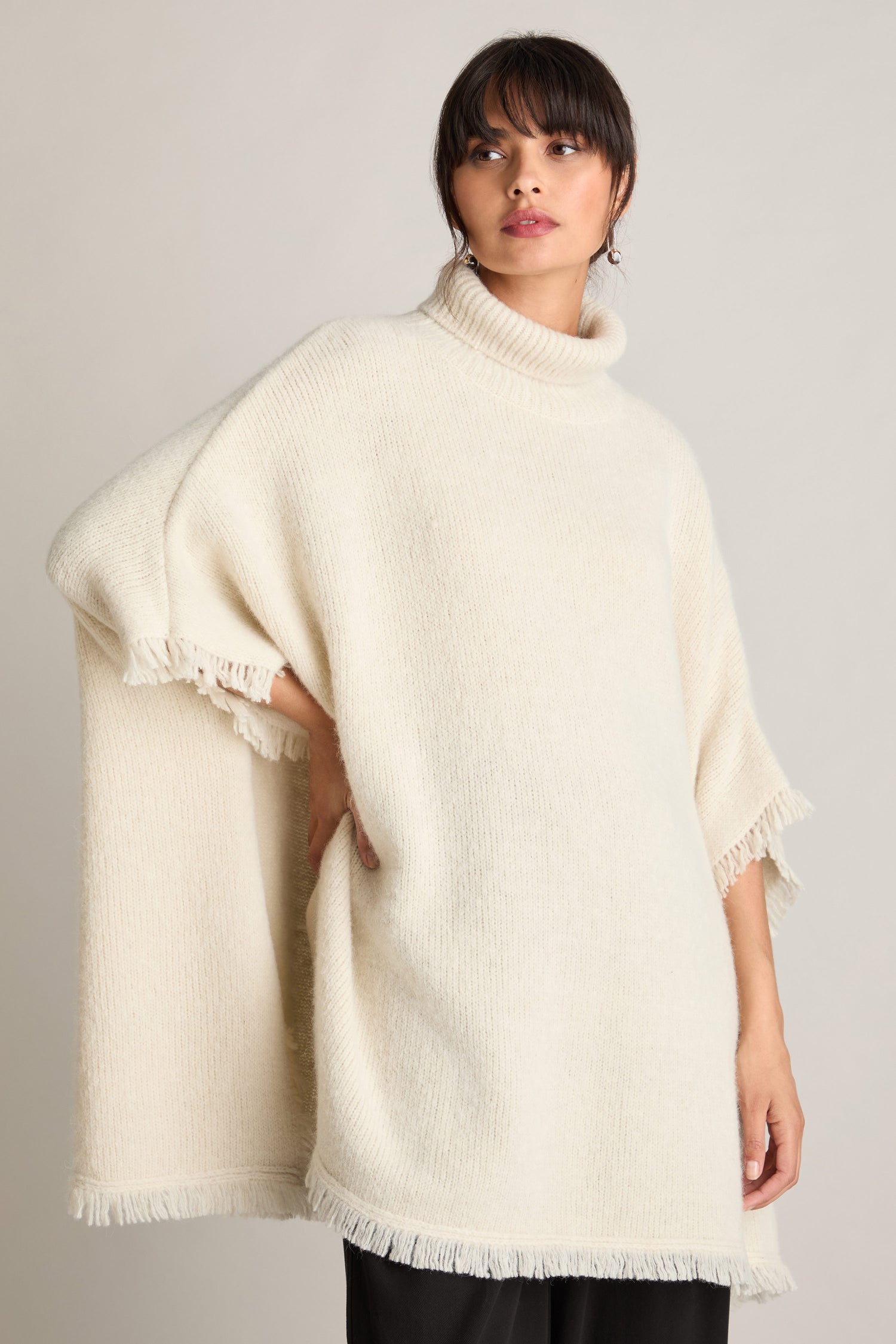 A person wearing a loose-fitting, cream-colored Cashmere And Alpaca Poncho featuring fringe details and a cozy polo neck stands against a plain background.