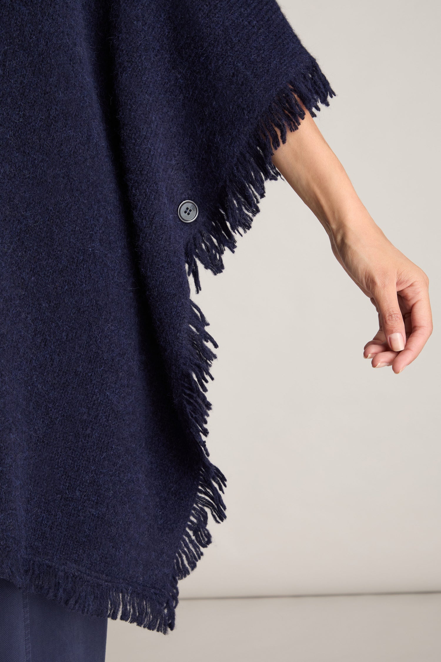 A close-up of a person wearing the luxurious and comfortable Cashmere And Alpaca Poncho in blue, adorned with fringes and button details, as they extend their right arm against a plain, neutral background.