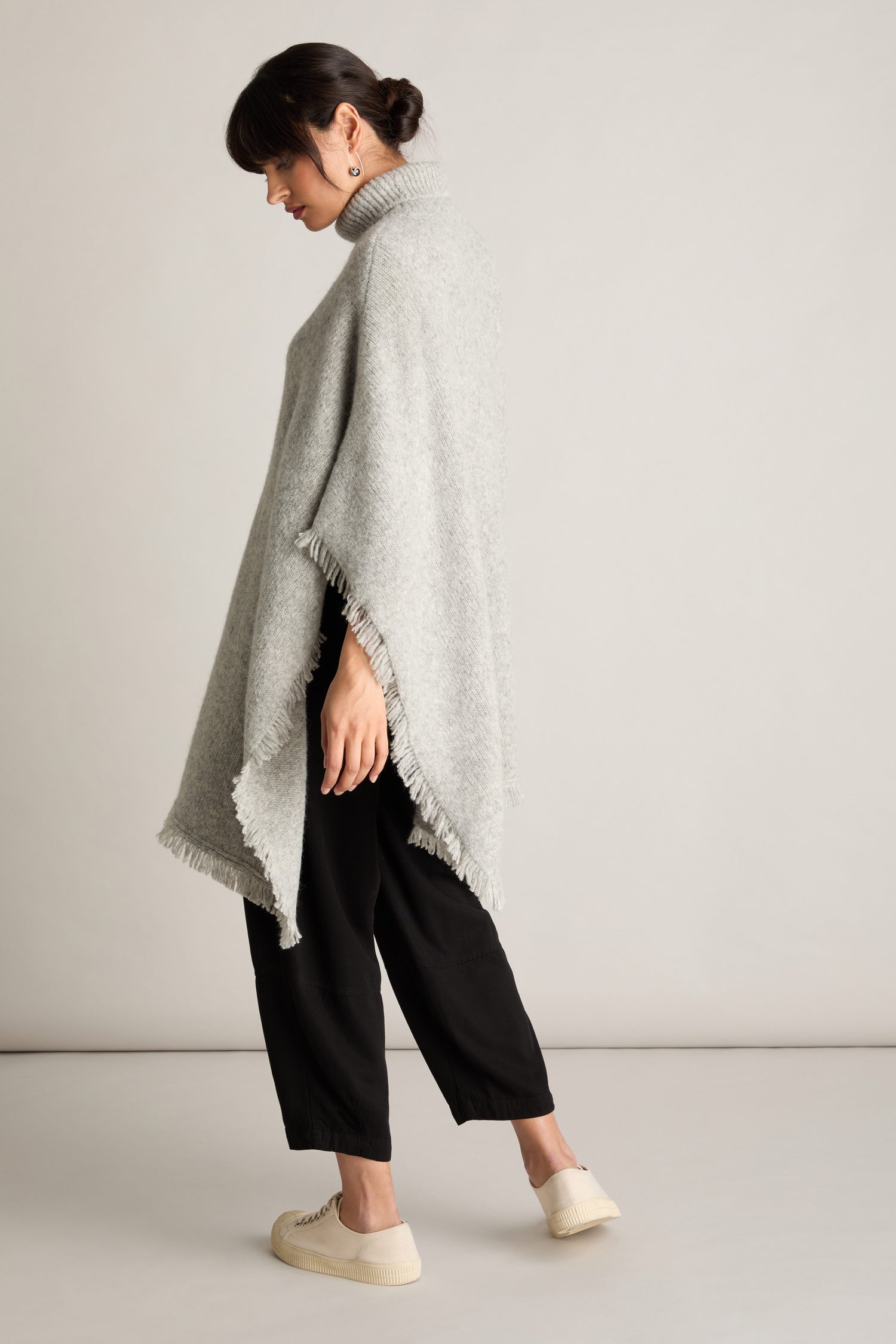 A person stands sideways against a plain background, exuding luxury and comfort in a light gray Cashmere And Alpaca Poncho with fringe detailing, paired with black pants and white sneakers.