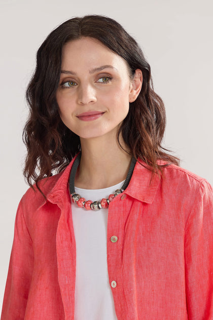 A woman with wavy, dark hair is wearing a coral-colored shirt over a white top and a Coral Bead Cord Necklace as her statement piece. She is looking slightly to her left and smiling gently.