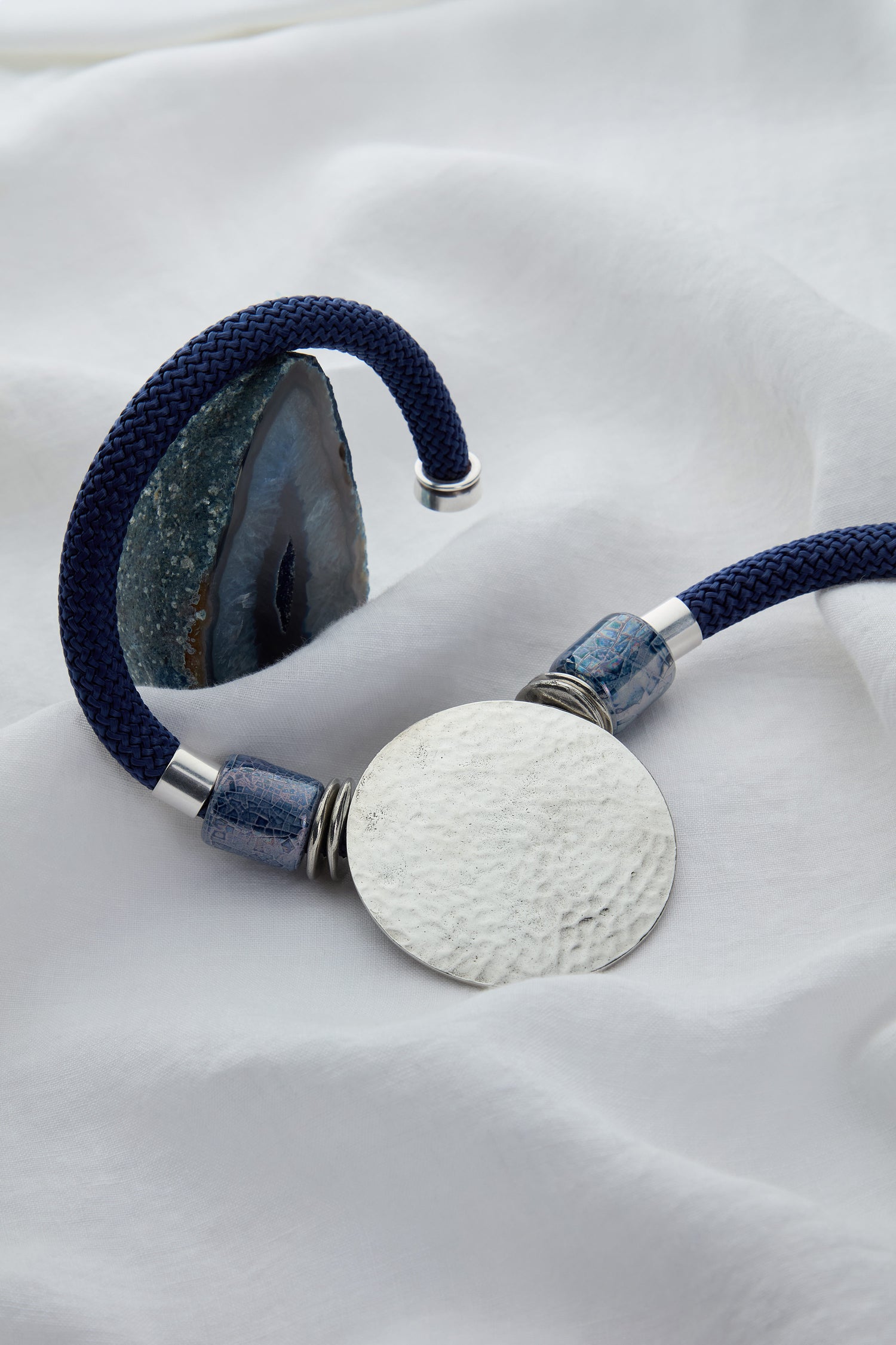 Image of a necklace with a blue braided cord, featuring a polished agate sphere on one end and a large textured silver disc on the other, placed on a white fabric background. The unique design echoes the elegance of Etnika jewellery.