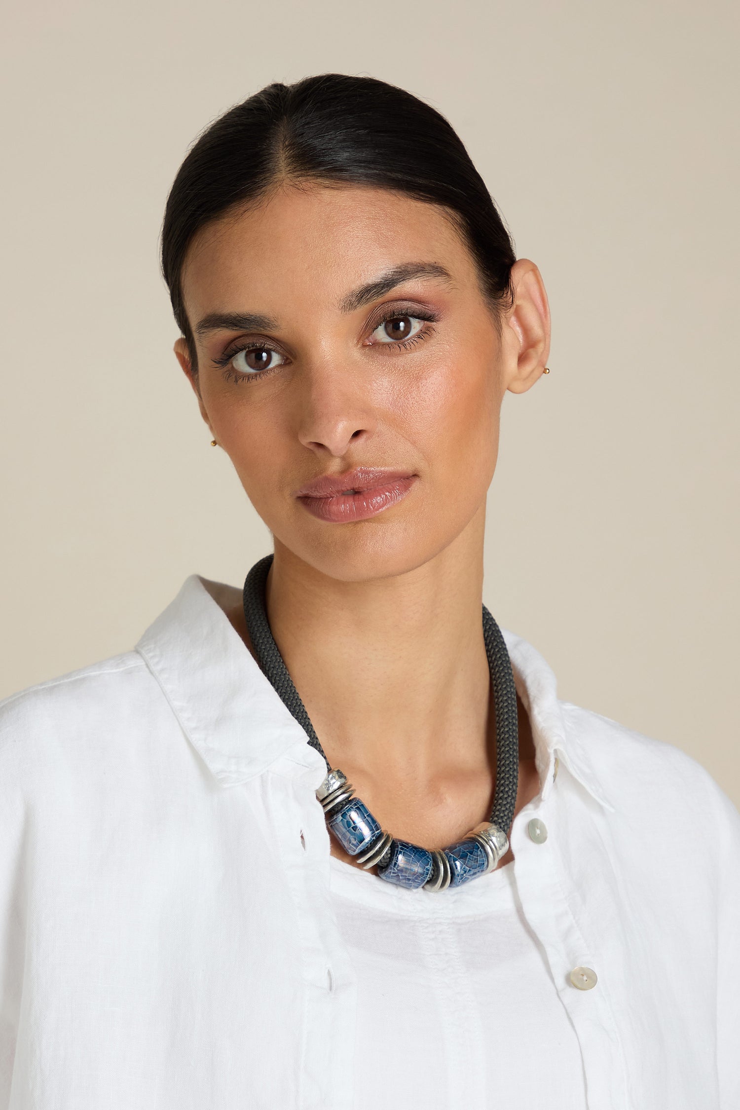 A person with dark hair tied back, wearing a white shirt and the Trio Bead Cord Necklace, stands against a neutral background, showcasing unique statement Art-lead jewellery.