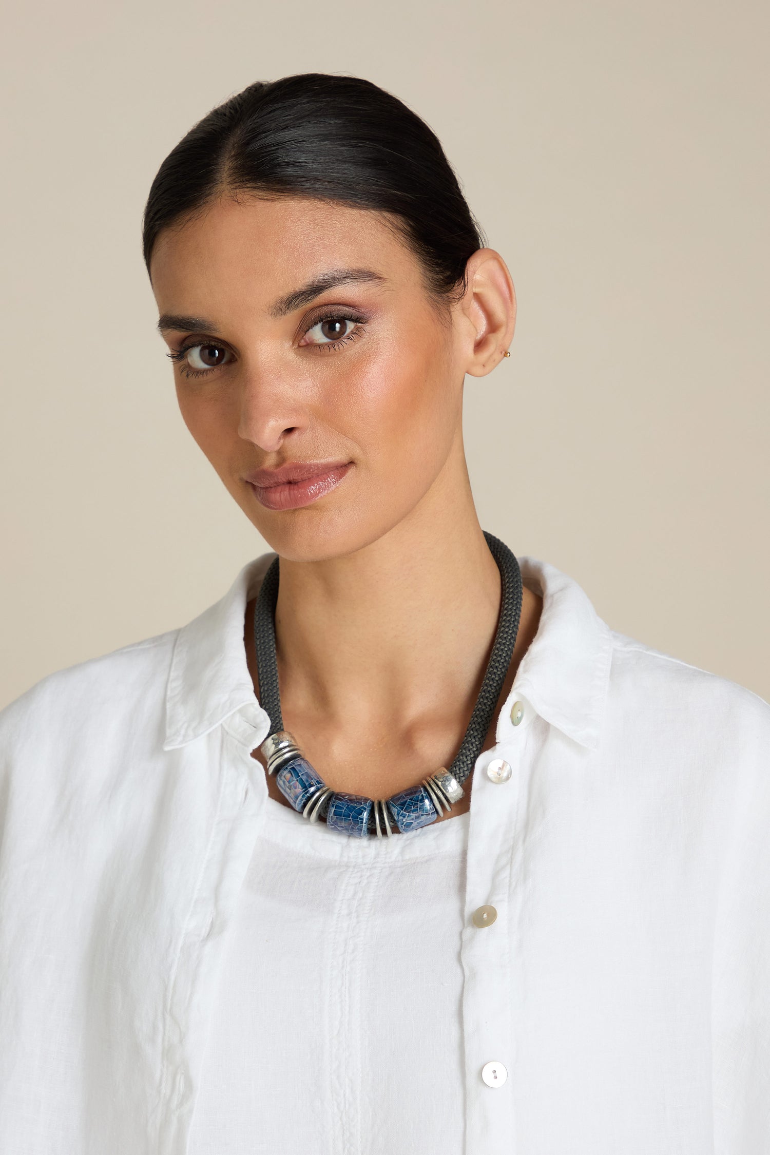 A woman with dark hair in an updo is wearing a white shirt and the stunning Trio Bead Cord Necklace, featuring blue and silver beads, against a beige background.