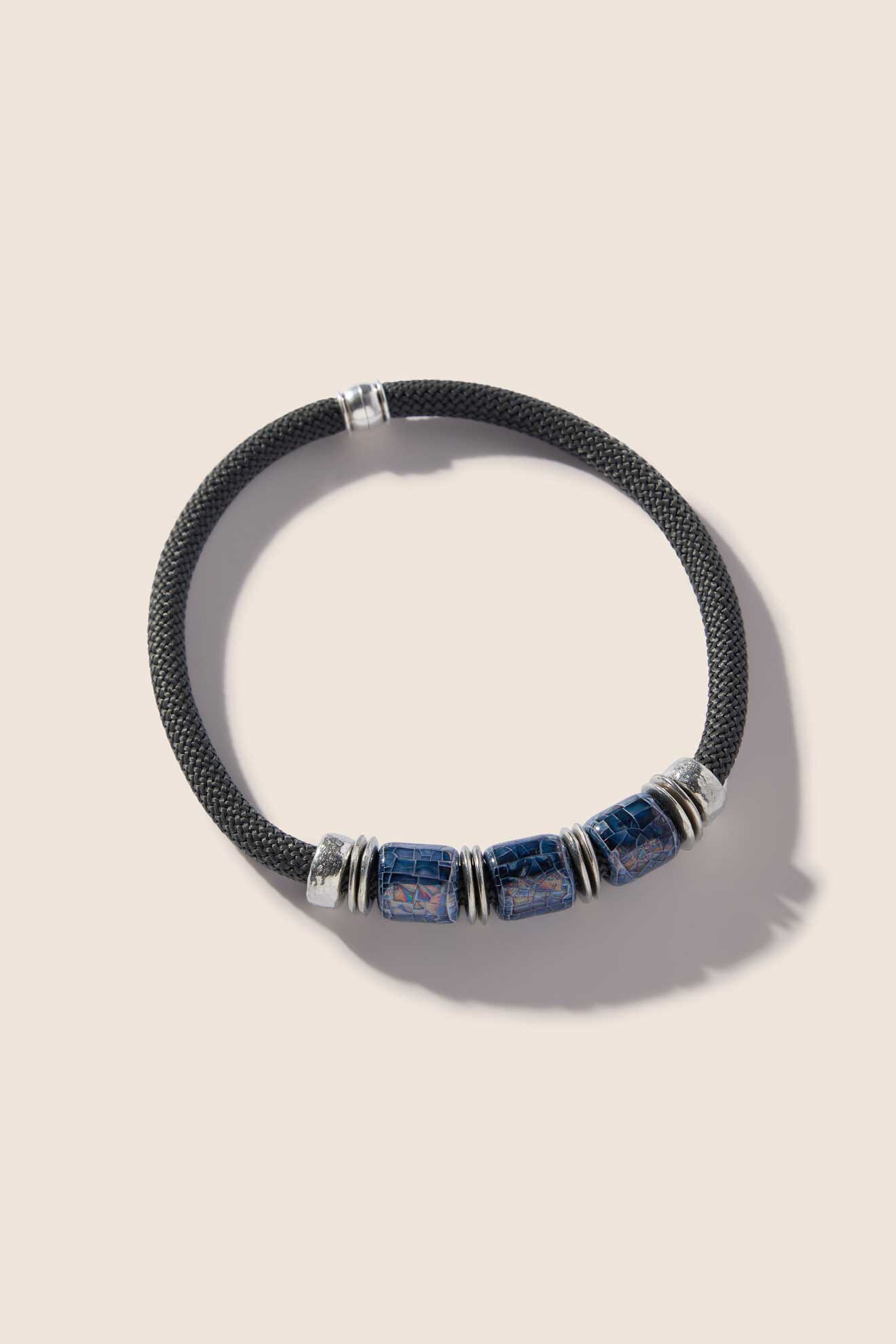 The Trio Bead Cord Necklace, a black woven creation featuring three blue marbled beads separated by silver rings, lies on a light background—a timeless design from our jewellery studio.