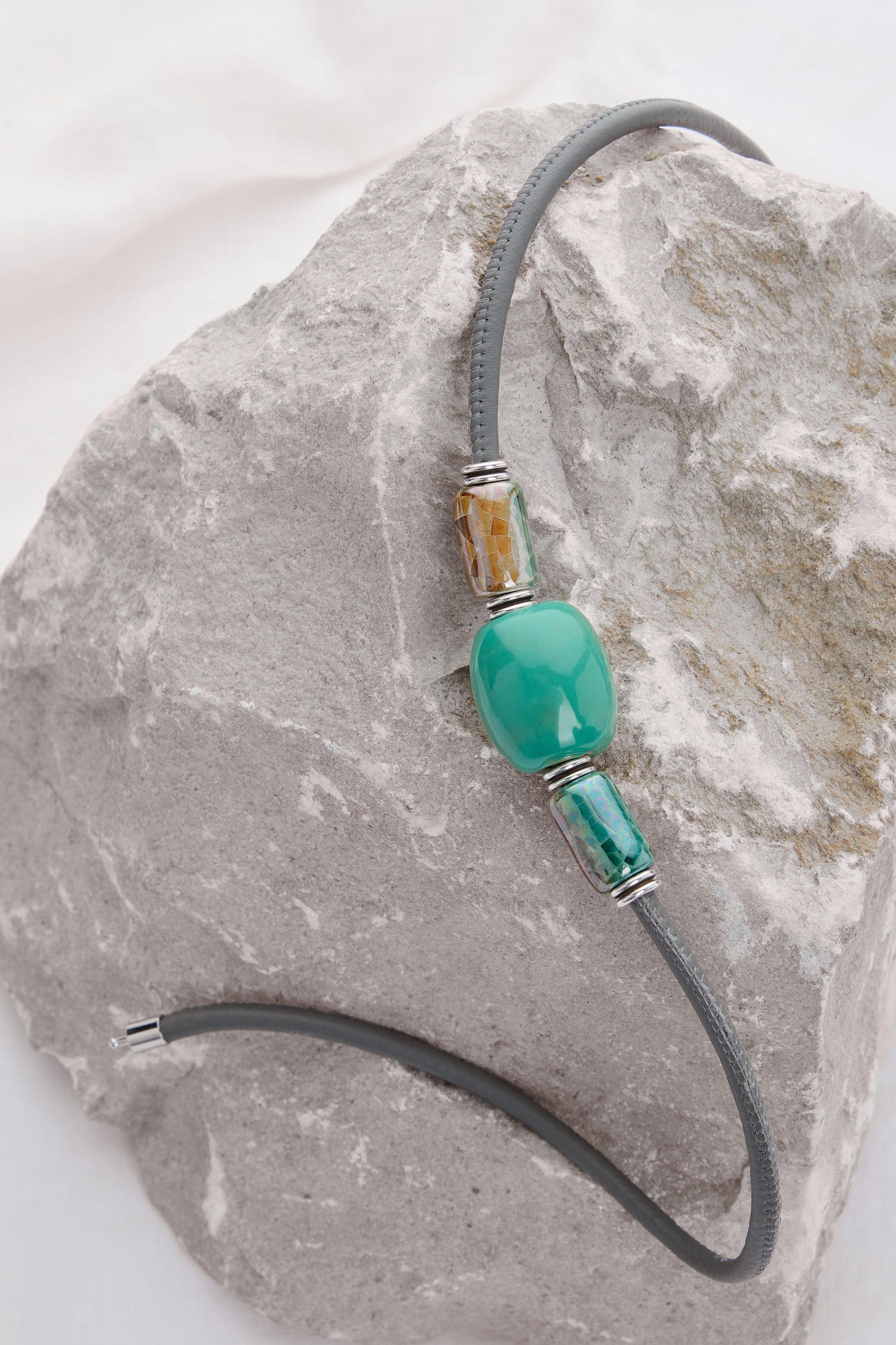 Gray cord necklace with Etnika's signature blue marbled beads, along with teal, green, and amber stones, placed on a rough light gray rock surface has been replaced with Triple Bead Leather Cord Necklace.