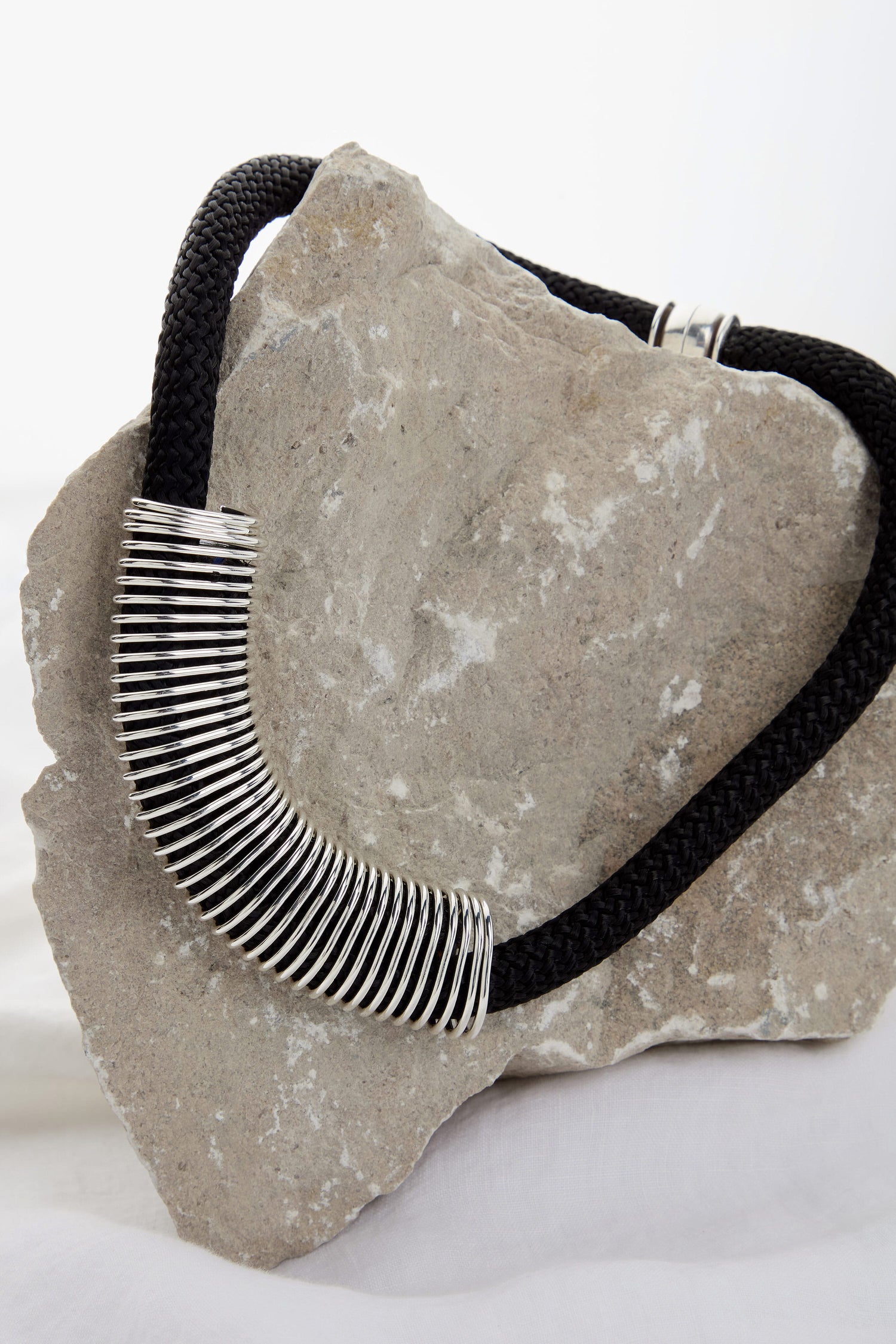 A Spring Cord Necklace adorned with a curved segment of silver rings is displayed draped over a large grey rock, featuring a magnetic closure for easy wear.