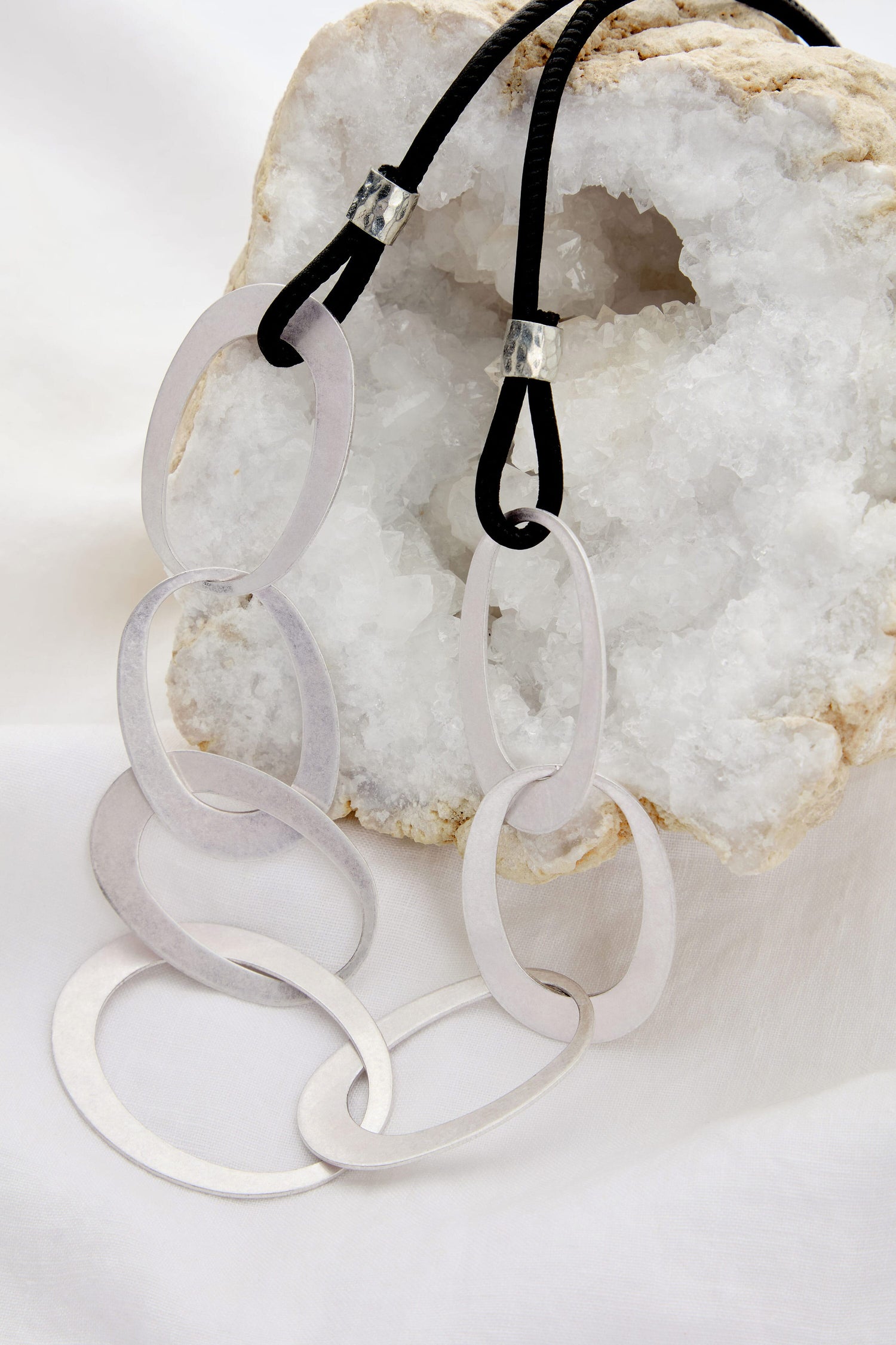 The minimalist Silver Links Leather Cord Necklace, featuring large silver oval links on a black leather cord, is elegantly displayed against a white geode rock and fabric backdrop.